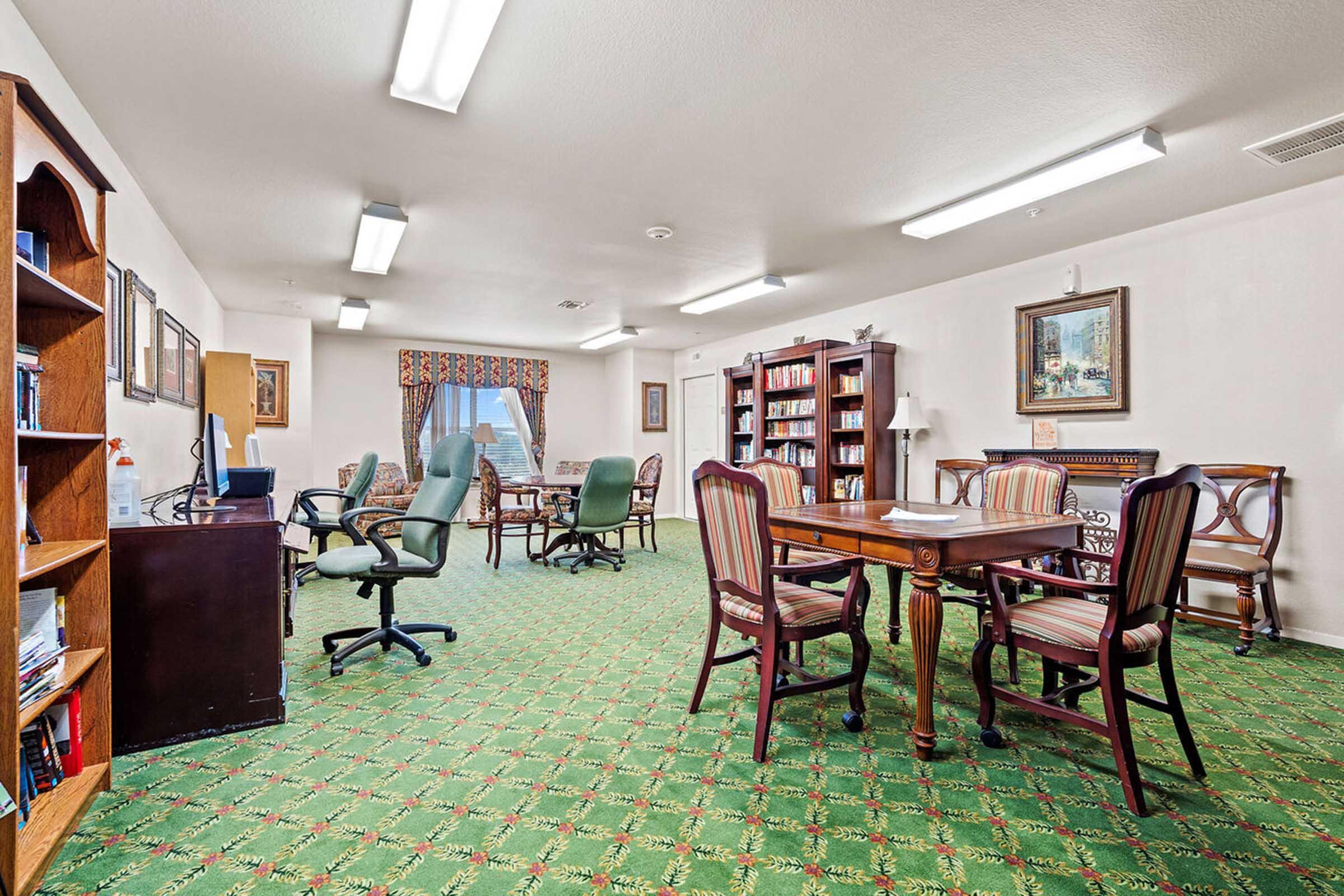 A spacious room with green carpet featuring floral patterns. There are bookshelves filled with books, a wooden table surrounded by chairs, and several comfortable seating areas.