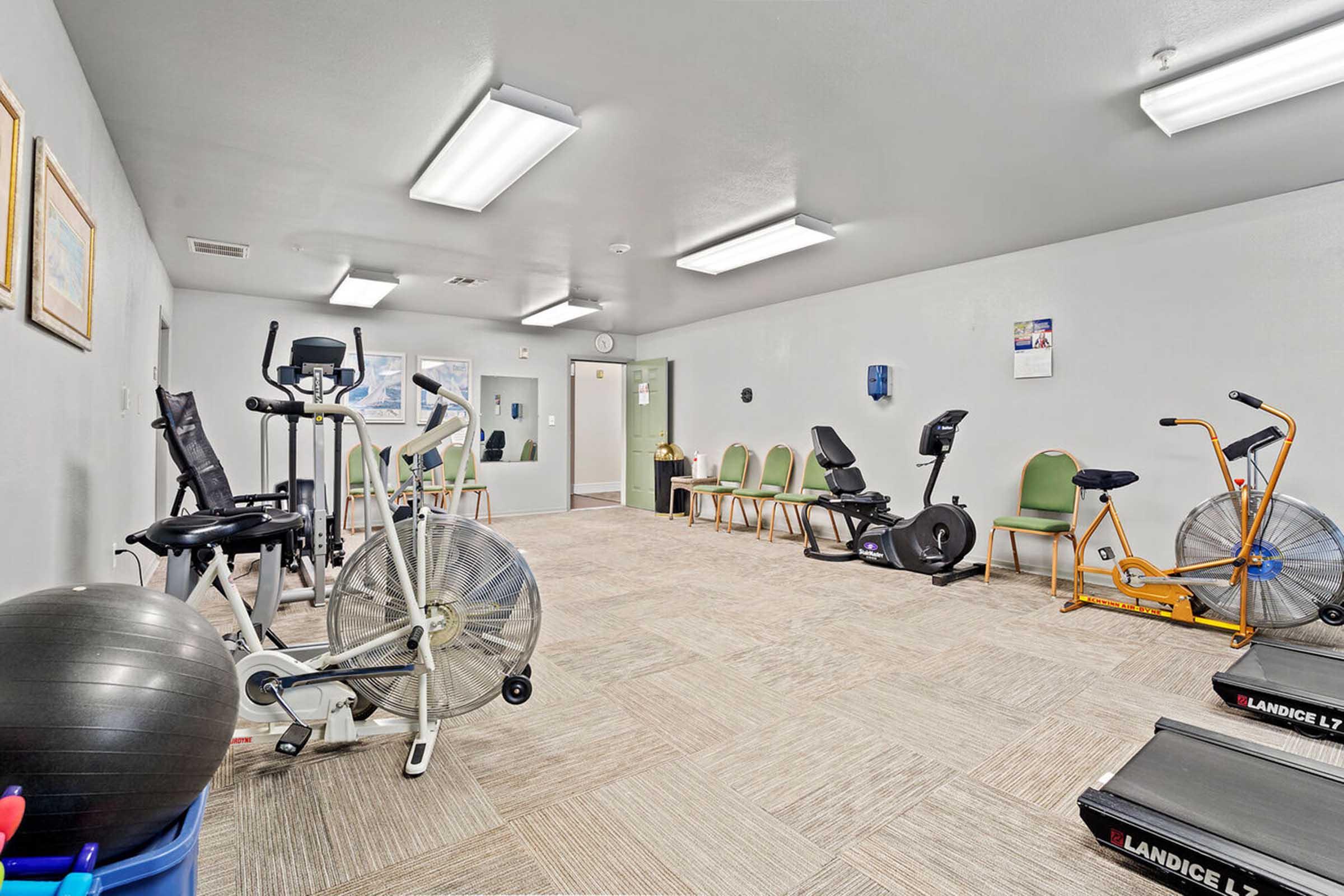 A spacious exercise room featuring various workout equipment, including stationary bikes, a fan bike, an exercise ball, and multiple chairs arranged along the wall.
