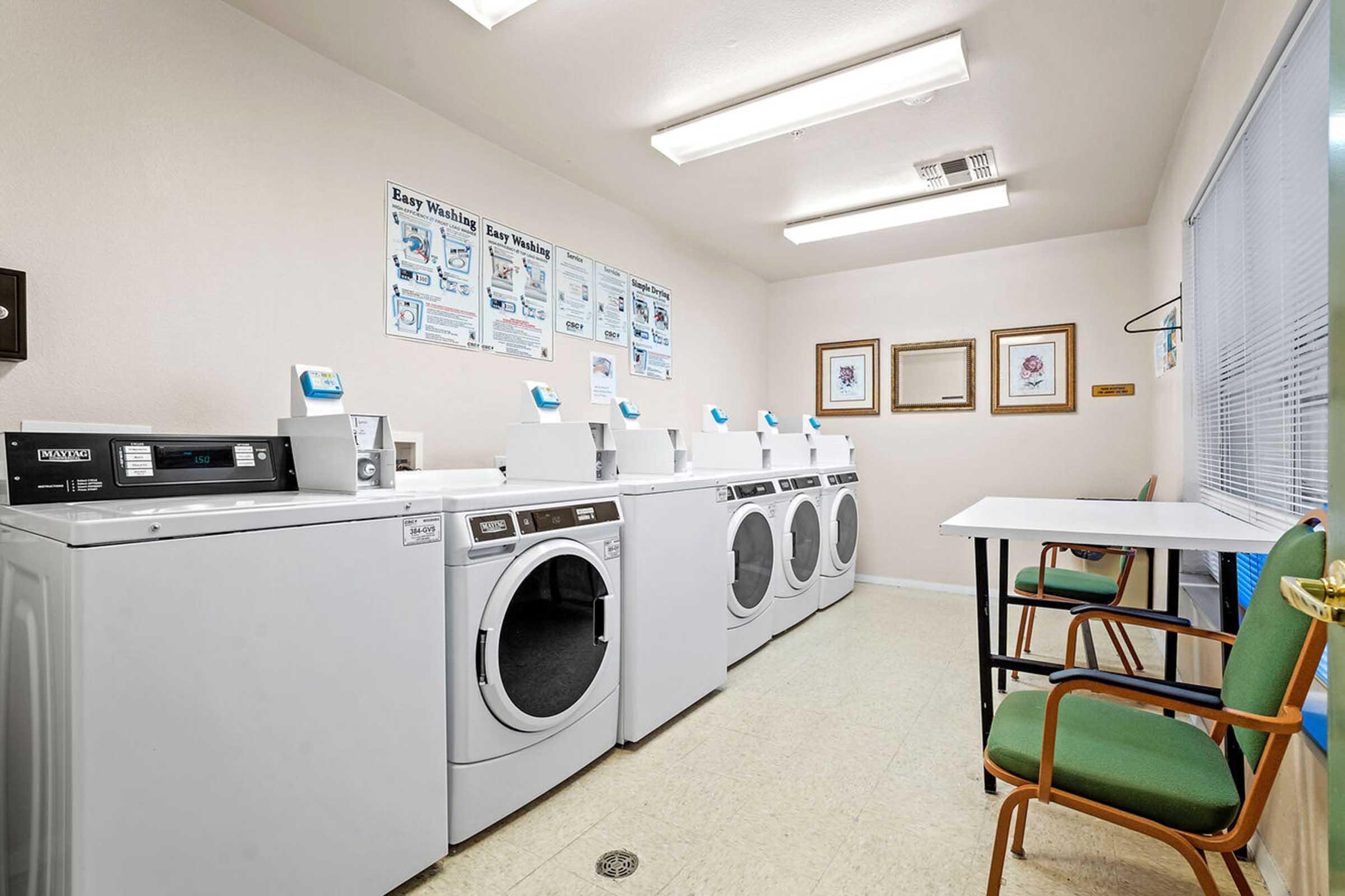 A clean and well-lit laundry room featuring several white washing machines and dryers, with a folding table and two green chairs.