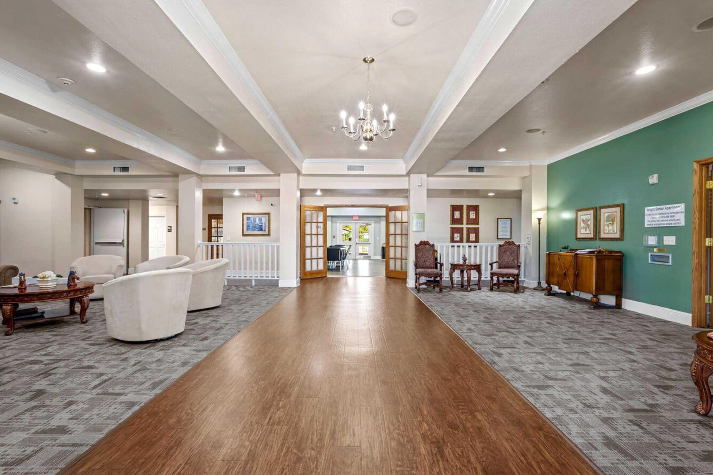 A spacious common area featuring a wooden floor, elegant chandelier, and ample seating with white armchairs and antique-style wooden furniture.
