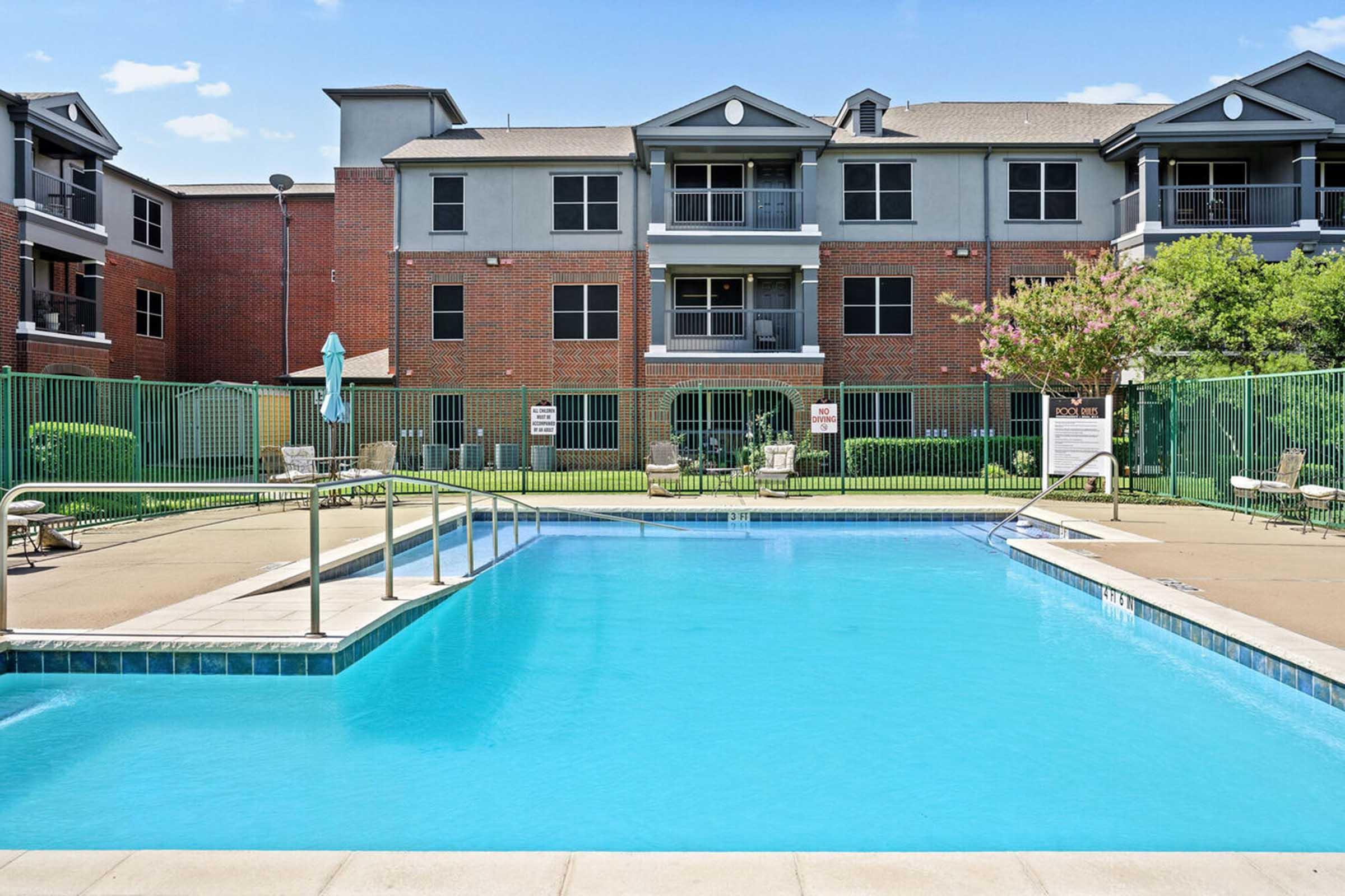 A clear swimming pool in foreground, surrounded by lounge chairs. In the background, a multi-story apartment building with a mix of brick and siding.