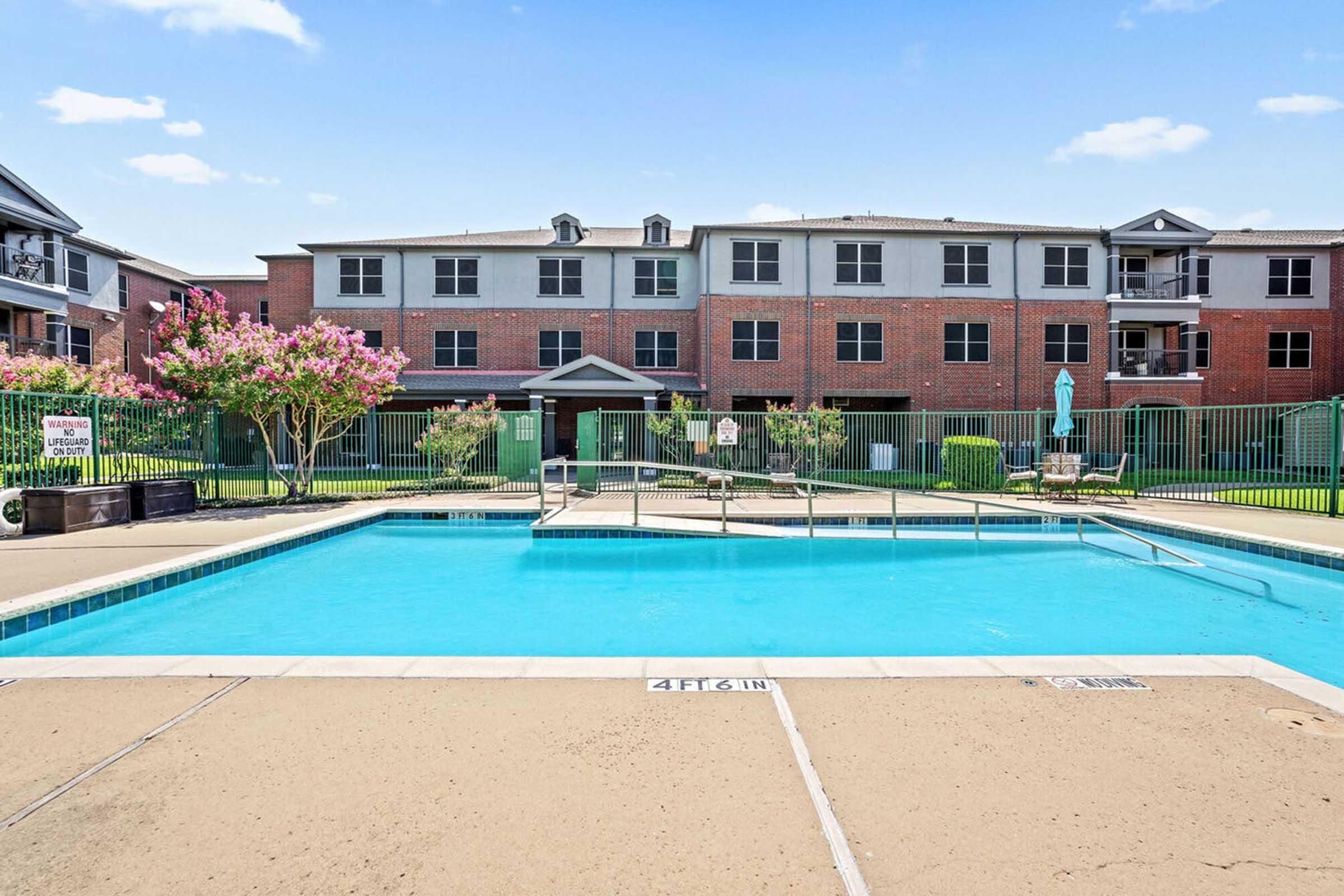 A clear swimming pool surrounded by a sandy area and green landscaping, with a brick apartment building in the background.