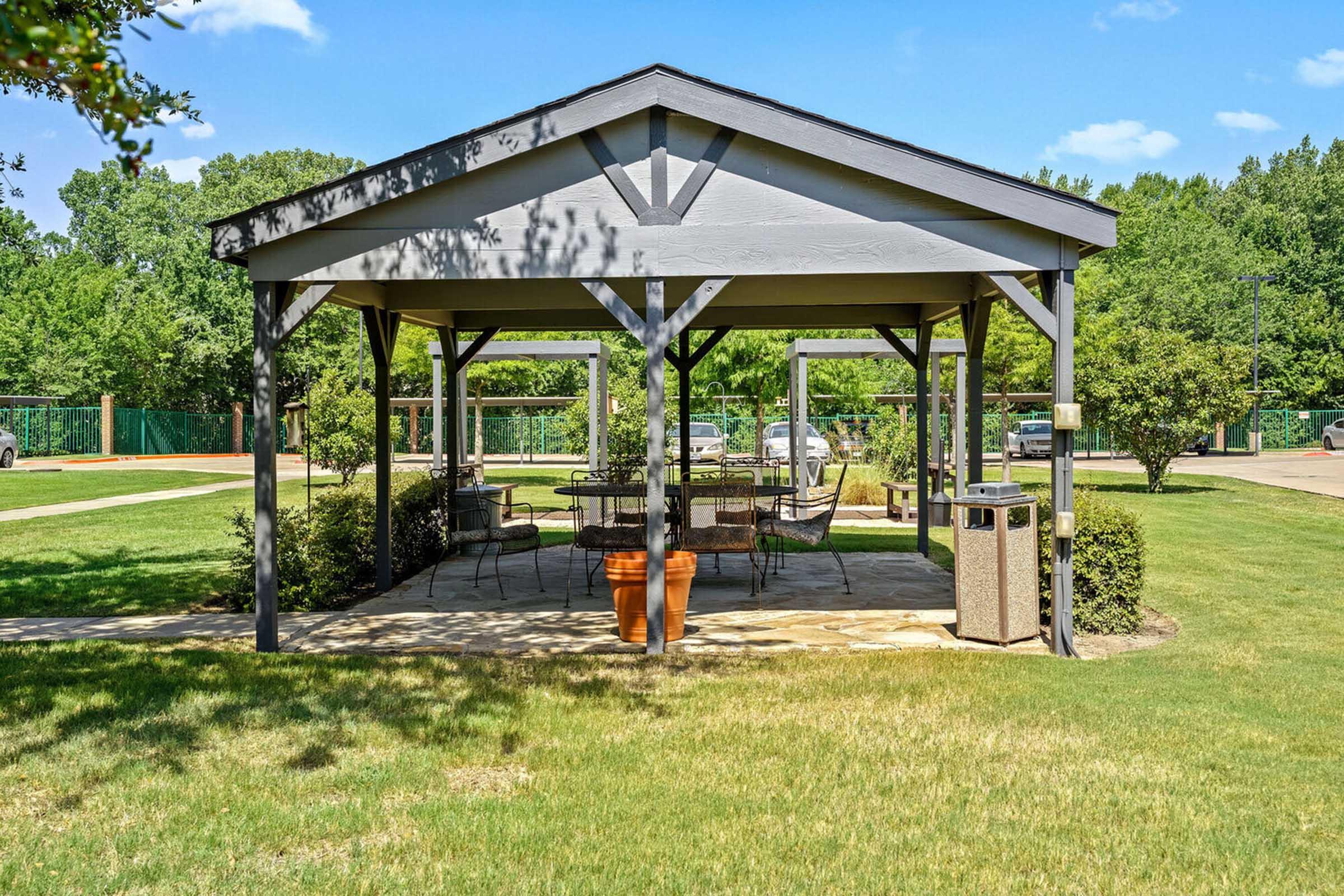 A shaded gazebo with a wooden frame and a stone floor, surrounded by green grass and shrubbery.
