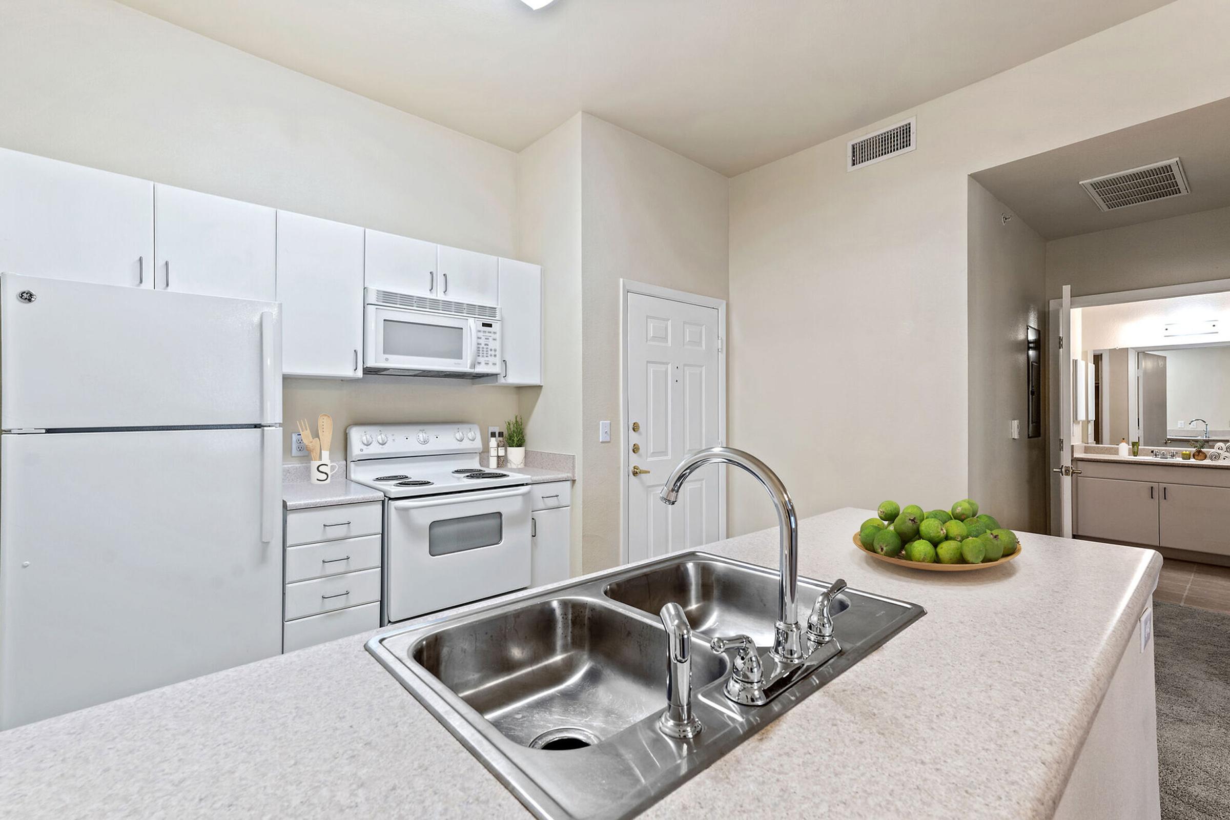 A modern kitchen featuring white cabinetry, a double sink, and a countertop with a bowl of green limes. There is a white refrigerator, microwave, and oven. The space is well-lit, with neutral-colored walls and an adjoining room visible in the background.