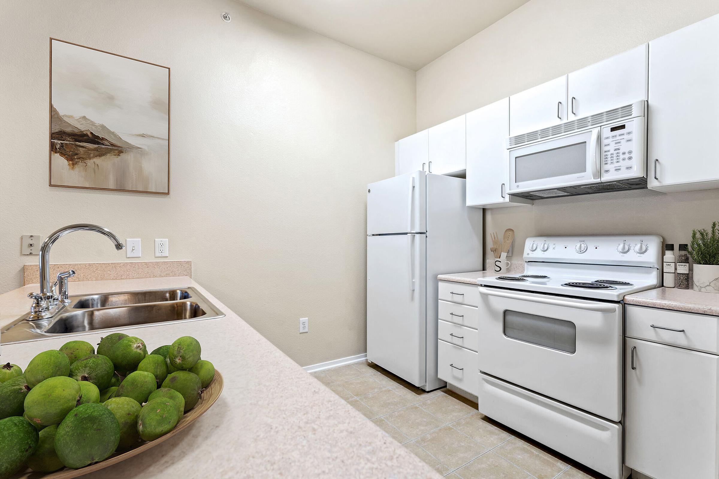 A bright kitchen featuring a white stove and microwave, a refrigerator, and light-colored cabinetry. The countertop has a bowl of green limes, and there’s a sink with a modern faucet. A piece of wall art hangs above the countertop, adding a subtle decorative touch. Neutral tones create a clean, inviting atmosphere.