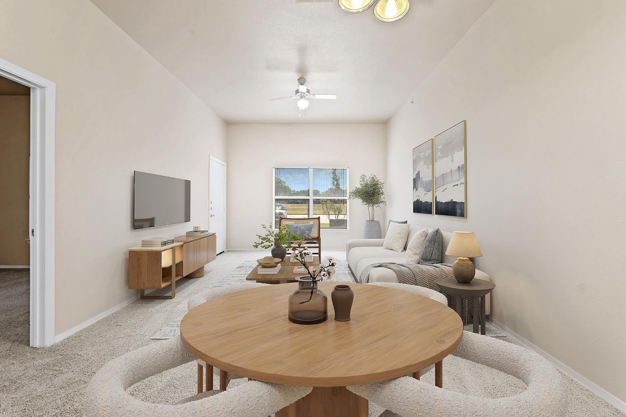 A well-lit living room featuring a cozy seating area with a light-colored sofa and a wooden coffee table. The decor includes a plant, a TV on a wooden console, and artwork on the walls. Large windows allow natural light to fill the space, creating a warm and inviting atmosphere.