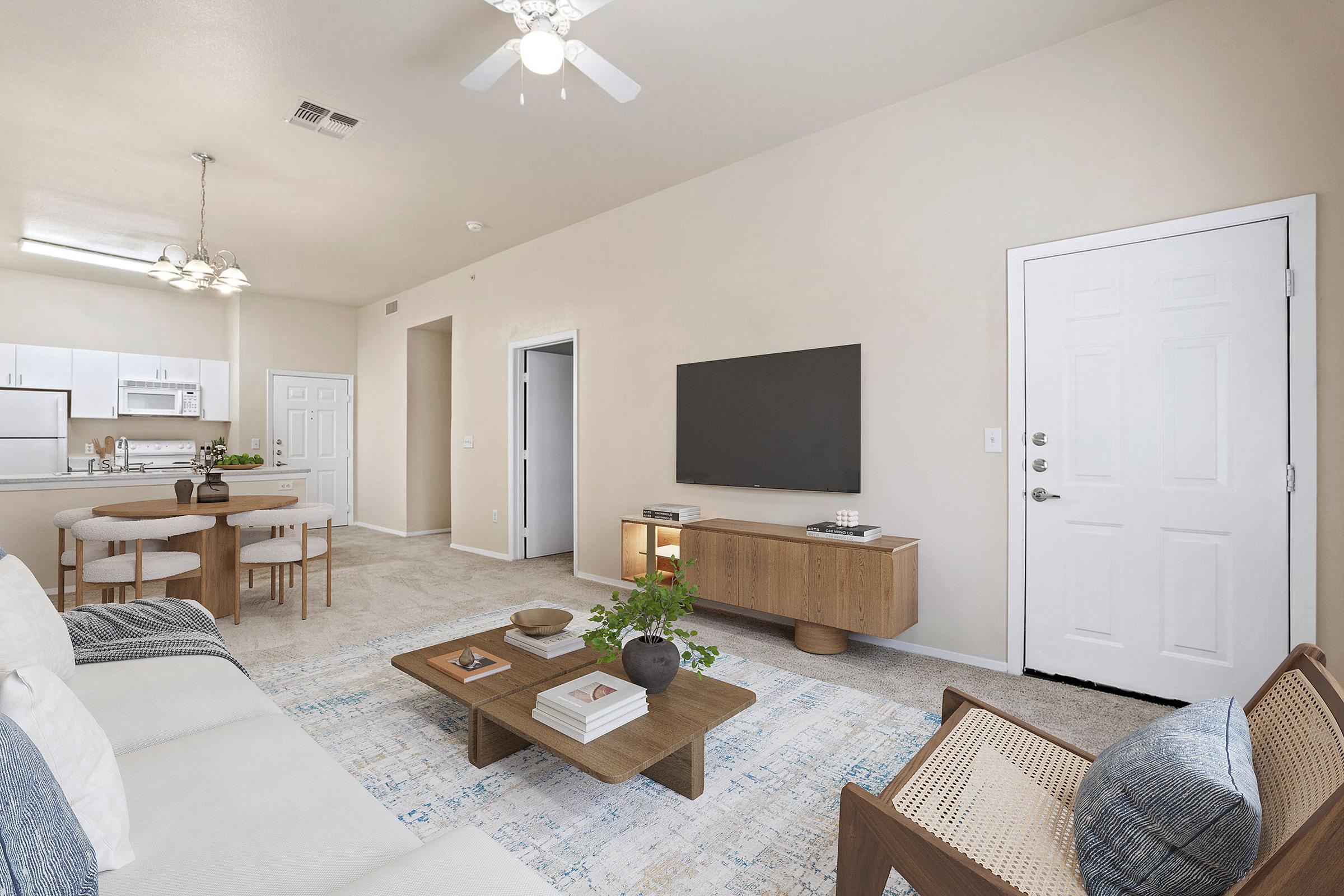 A modern living room with light beige walls and carpet, featuring a white sofa, wooden furniture, a coffee table with decorative items, a large wall-mounted TV, and a round dining table. A door leads outside, and natural light fills the space, creating a welcoming atmosphere.