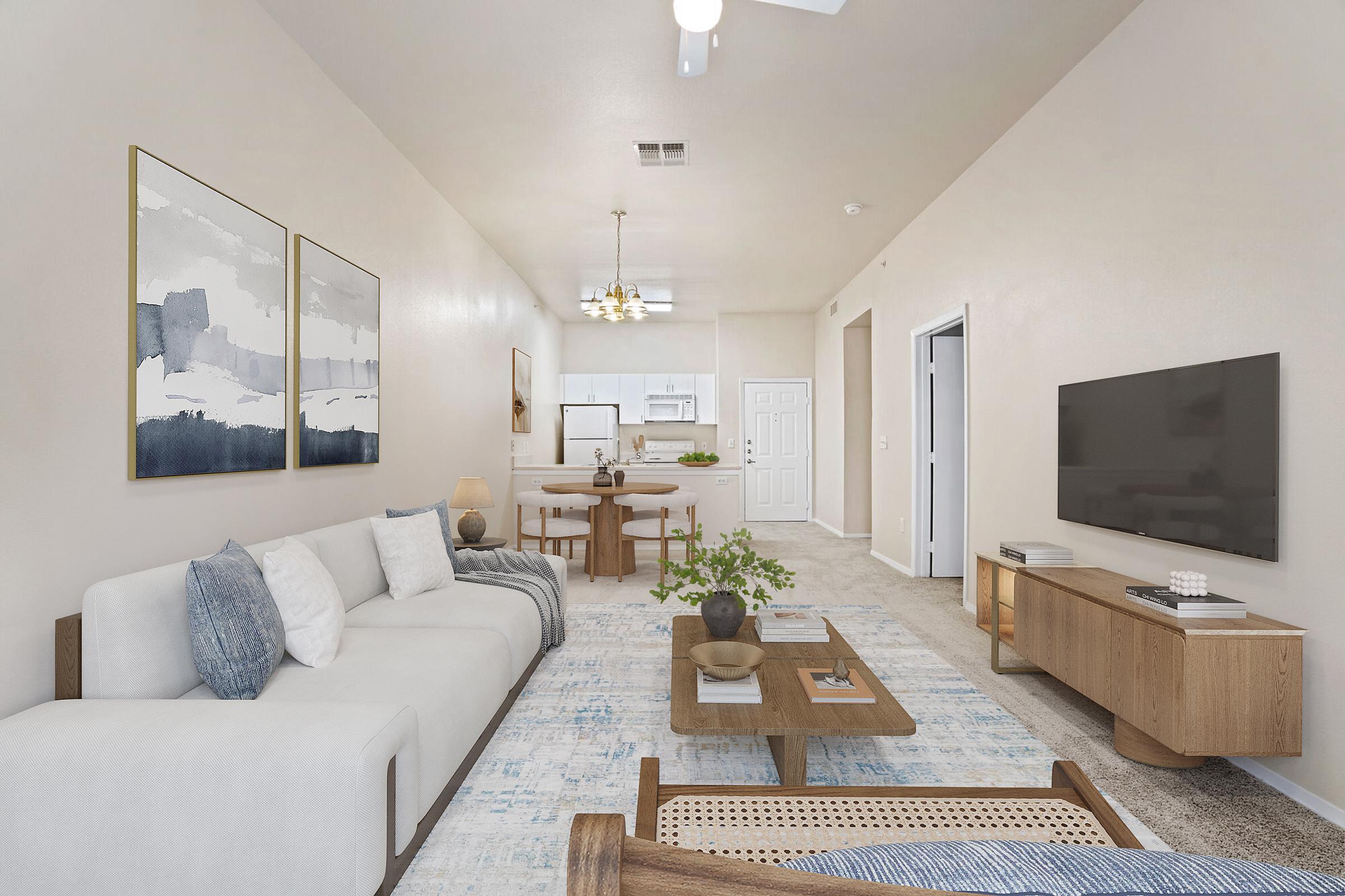 A bright and contemporary living room featuring a light-colored sofa, a wooden coffee table, and modern art on the walls. There's a television mounted on the wall opposite the sofa, and a dining area visible in the background. The room is well-lit, with a ceiling fan and decorative plants.