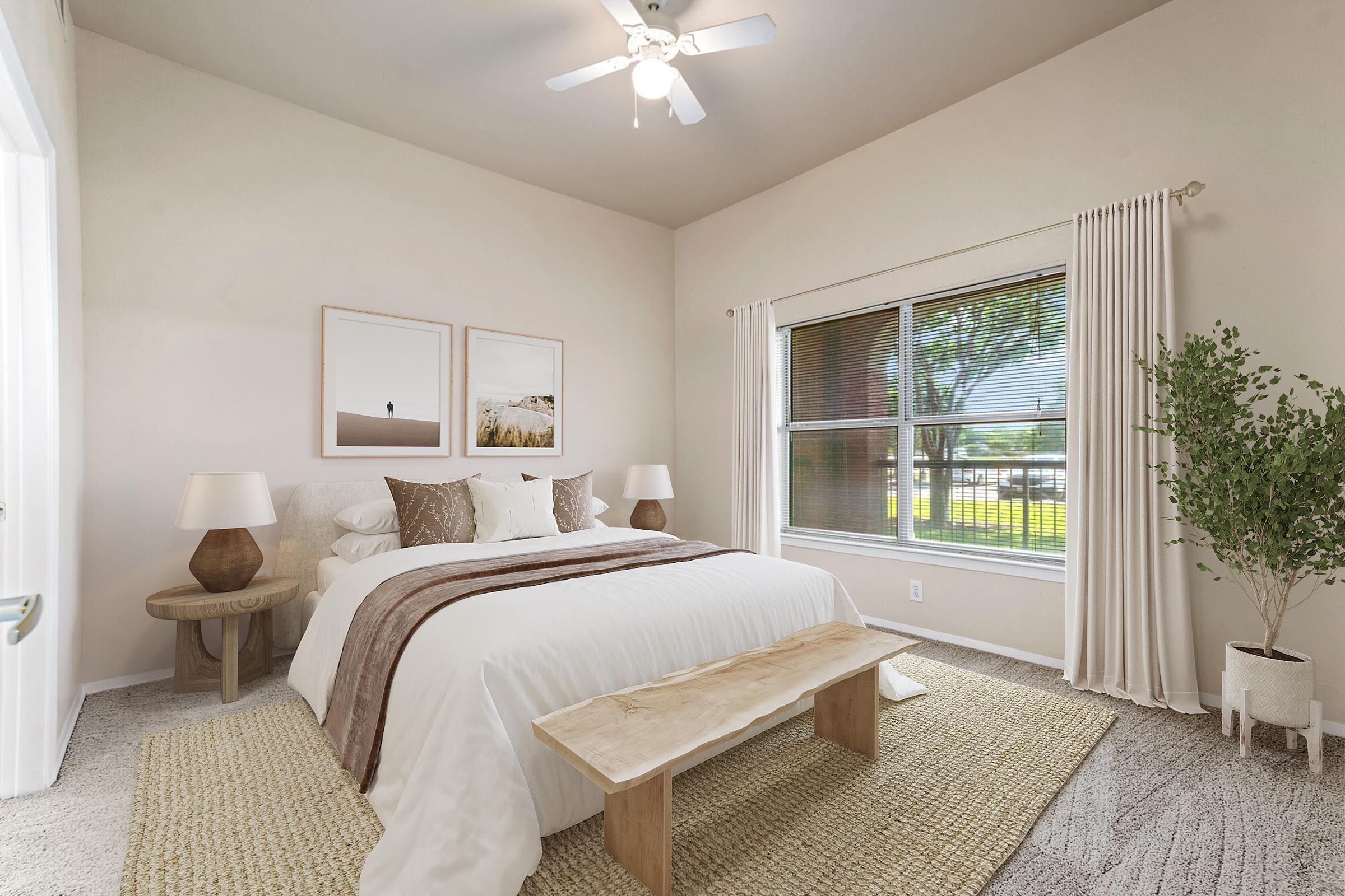 A cozy, well-lit bedroom featuring a large bed with white bedding, decorative pillows, and a throw blanket. Two art prints hang above the bed. There are light-colored bedside lamps, a wooden bench at the foot of the bed, and a textured area rug. A window with sheer curtains overlooks greenery outside.
