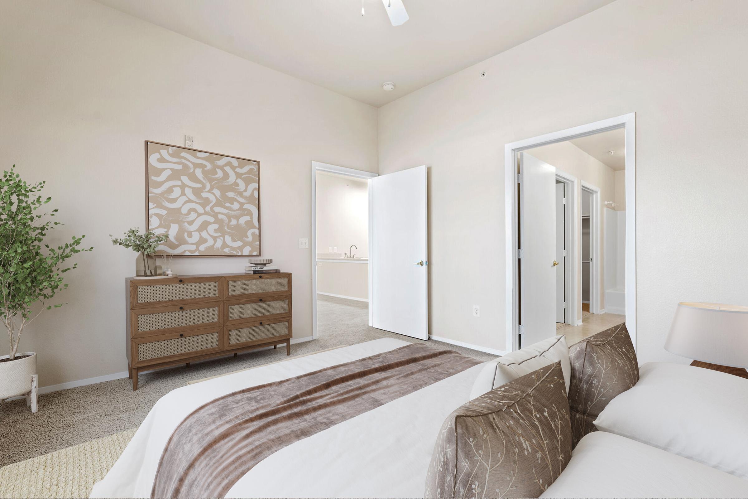 A bright and spacious bedroom with neutral wall colors, featuring a neatly made bed adorned with decorative pillows. A modern dresser stands against one wall, and there are open doors leading to other rooms. A plant adds a touch of greenery, while the lighting creates a warm and inviting atmosphere.