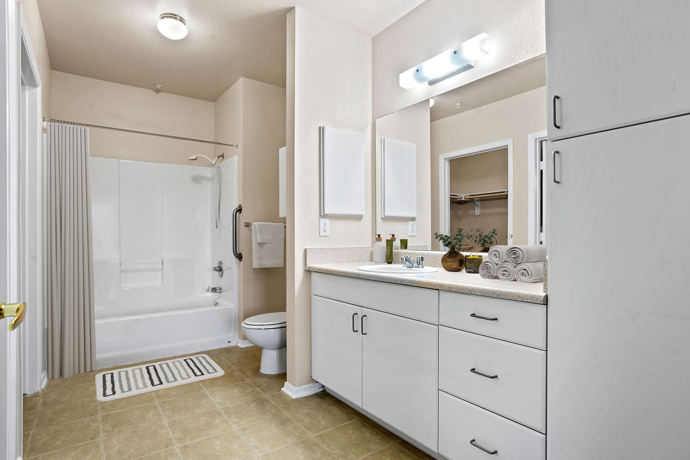 A well-lit bathroom featuring a white bathtub with a shower curtain, a toilet, and a large mirror over a vanity with a sink. The vanity has storage drawers and is adorned with decorative items, including towels and a plant. The floor is a neutral tile, and there's a small rug in front of the tub.