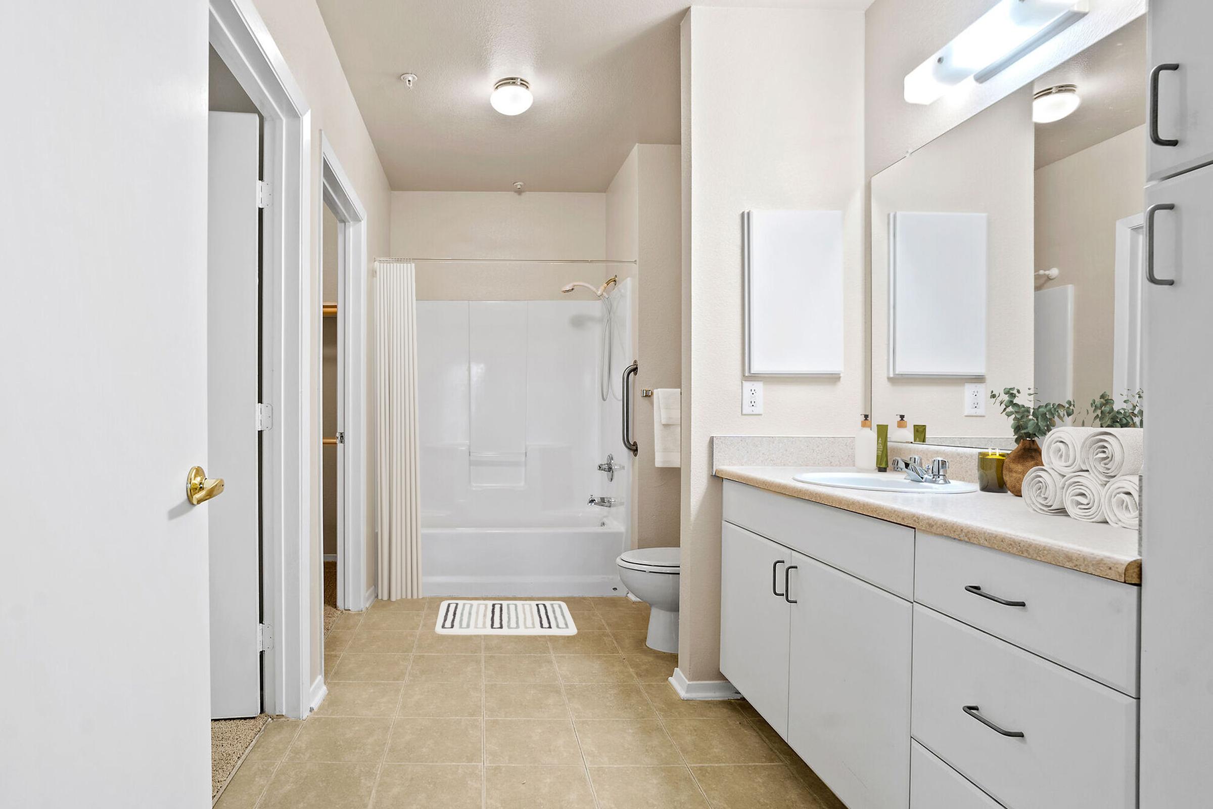 Modern bathroom featuring a tub/shower combination, a toilet, and dual sinks with a countertop. The space is well-lit with a mirror above each sink. Towel rolls are neatly arranged, and there are decorative elements on the counter. The flooring is tiled, and the walls are painted in neutral colors, creating a clean, inviting atmosphere.