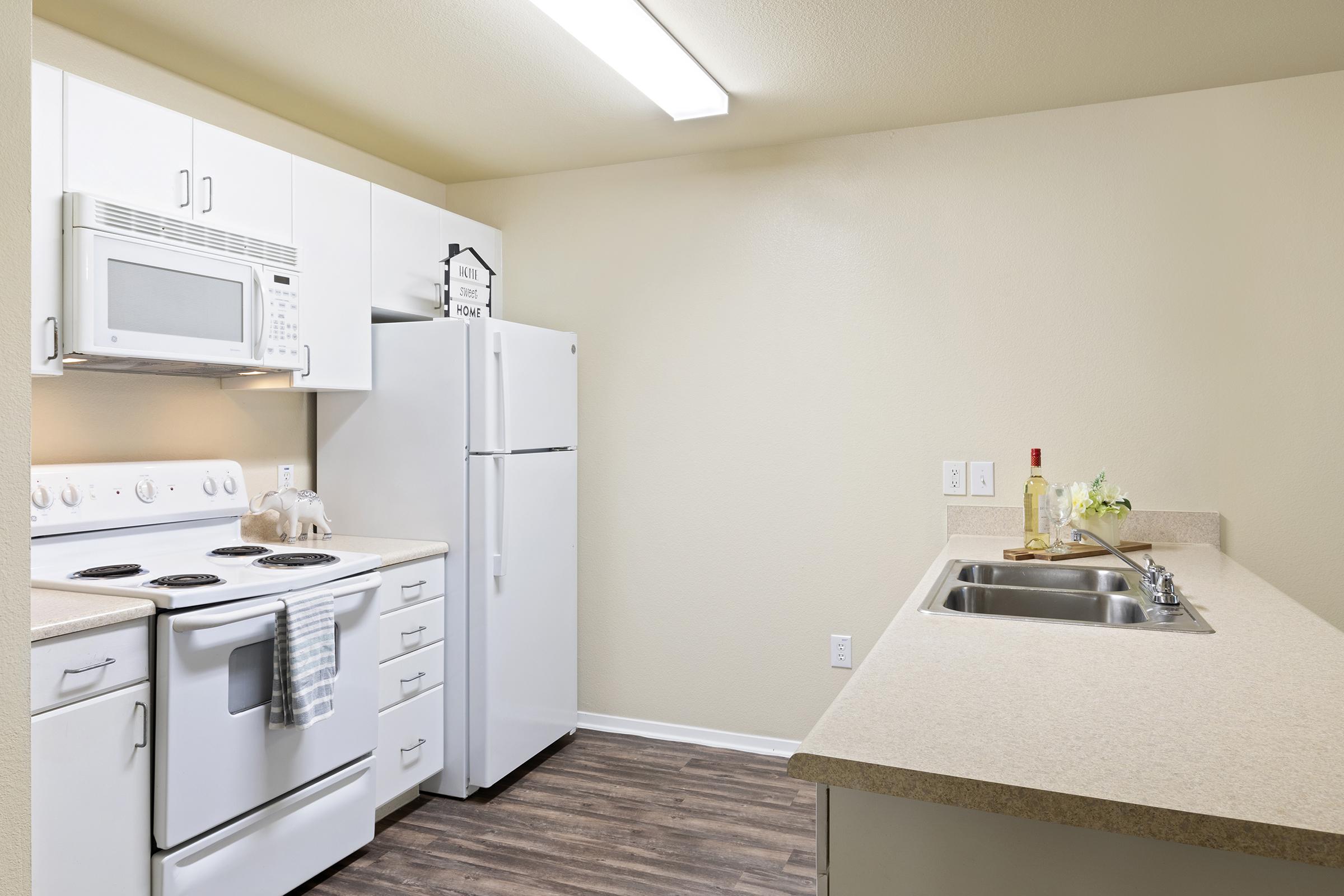 A modern kitchen featuring white cabinets and appliances, including a microwave, stove, and refrigerator. The countertops are light beige, and there are two sinks. A bottle of olive oil and a small plant are displayed on the counter, with light-colored walls and wooden flooring.