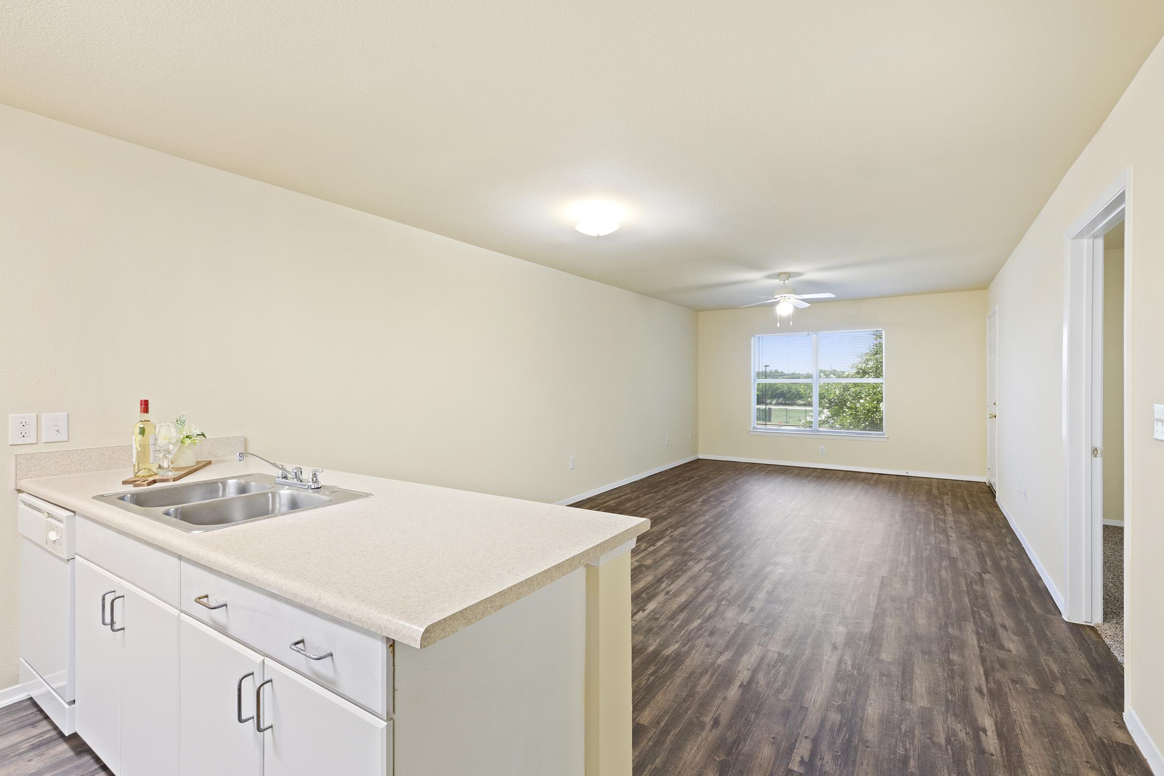 Bright and spacious living area featuring a kitchen with a sink and countertop on the left. The room has light-colored walls, a ceiling fan, and a large window allowing natural light. The flooring is dark wood, creating a warm contrast with the walls. An adjoining room is visible in the background.