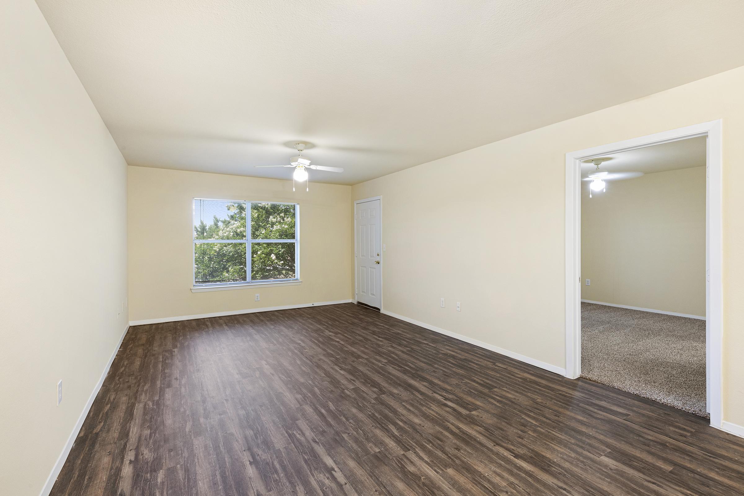 Spacious empty room with light-colored walls and a ceiling fan. A window lets in natural light, and there's a doorway leading to another room. The flooring appears to be a wood laminate style, and the overall atmosphere is bright and airy.