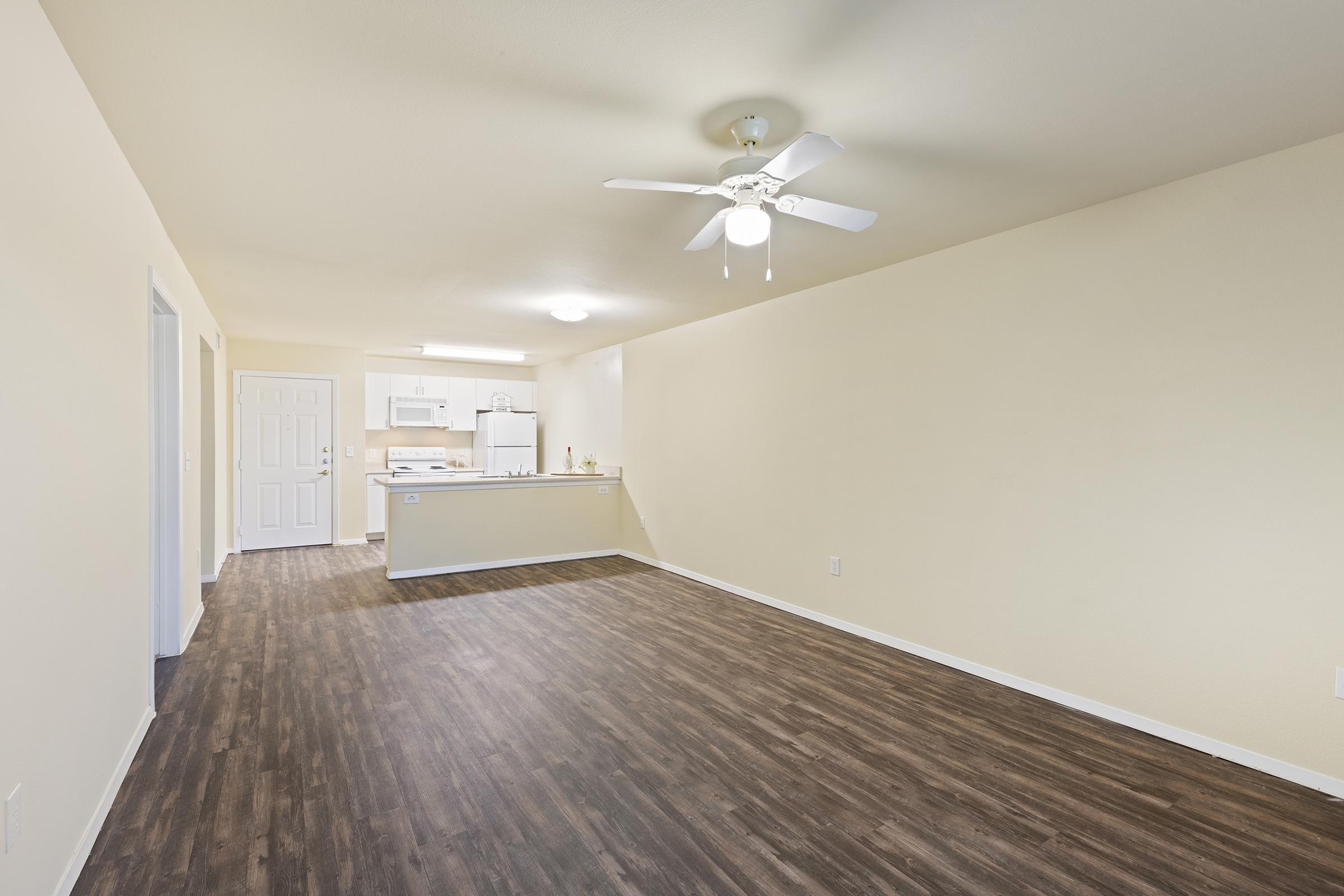 A spacious, empty living area with a light-colored wall and wooden laminate flooring. There is a ceiling fan overhead, and a kitchen can be seen in the background, featuring white appliances and cabinets. The room is well-lit, creating a bright and open atmosphere.