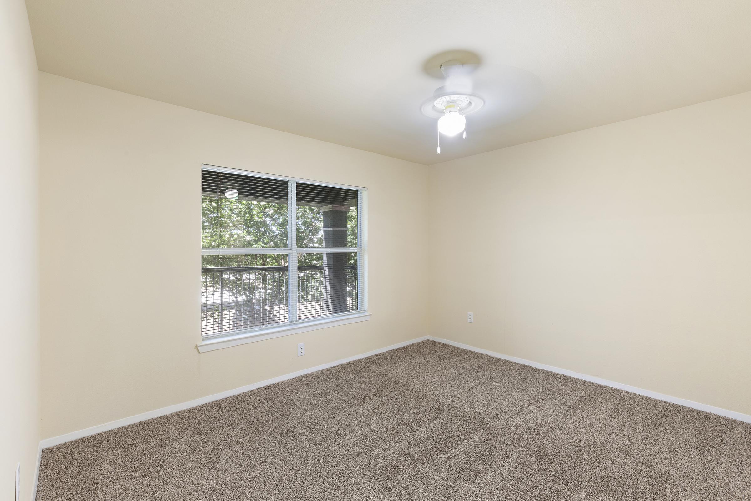 A clean, empty room with light beige walls and a ceiling fan. The floor is covered in light brown carpeting. A large window with blinds lets in natural light, showcasing a view of the outdoors. The space has a bright and airy feel, ideal for various uses such as a bedroom or office.