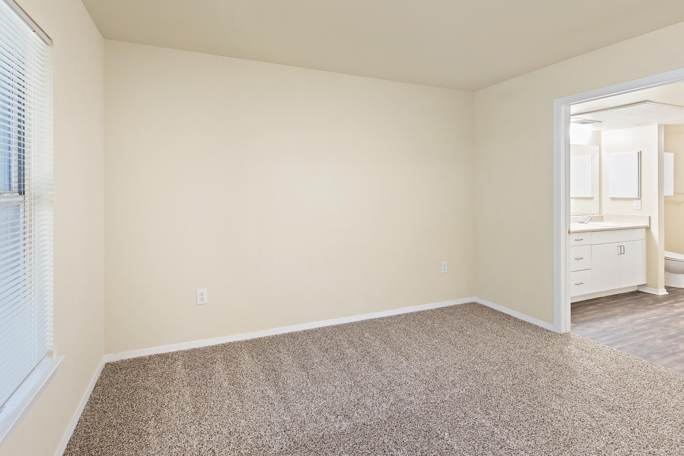 Empty room with beige walls and carpeted floor. A doorway on the right leads to a bathroom with white cabinetry and mirrors visible. Natural light comes in through a window on the left. The space is clean and unadorned, suitable for various uses or furnishings.
