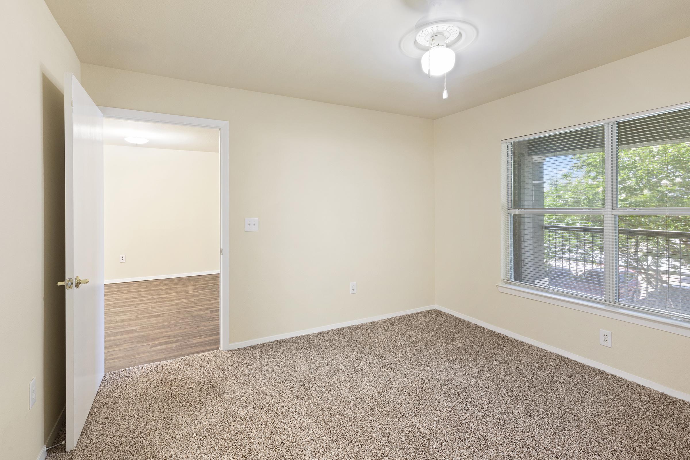 A vacant room featuring neutral beige walls and carpeted flooring. A ceiling fan is visible above, and a window allows natural light to enter. The room is sparsely decorated, with a door leading to another space in the background, emphasizing its open layout.