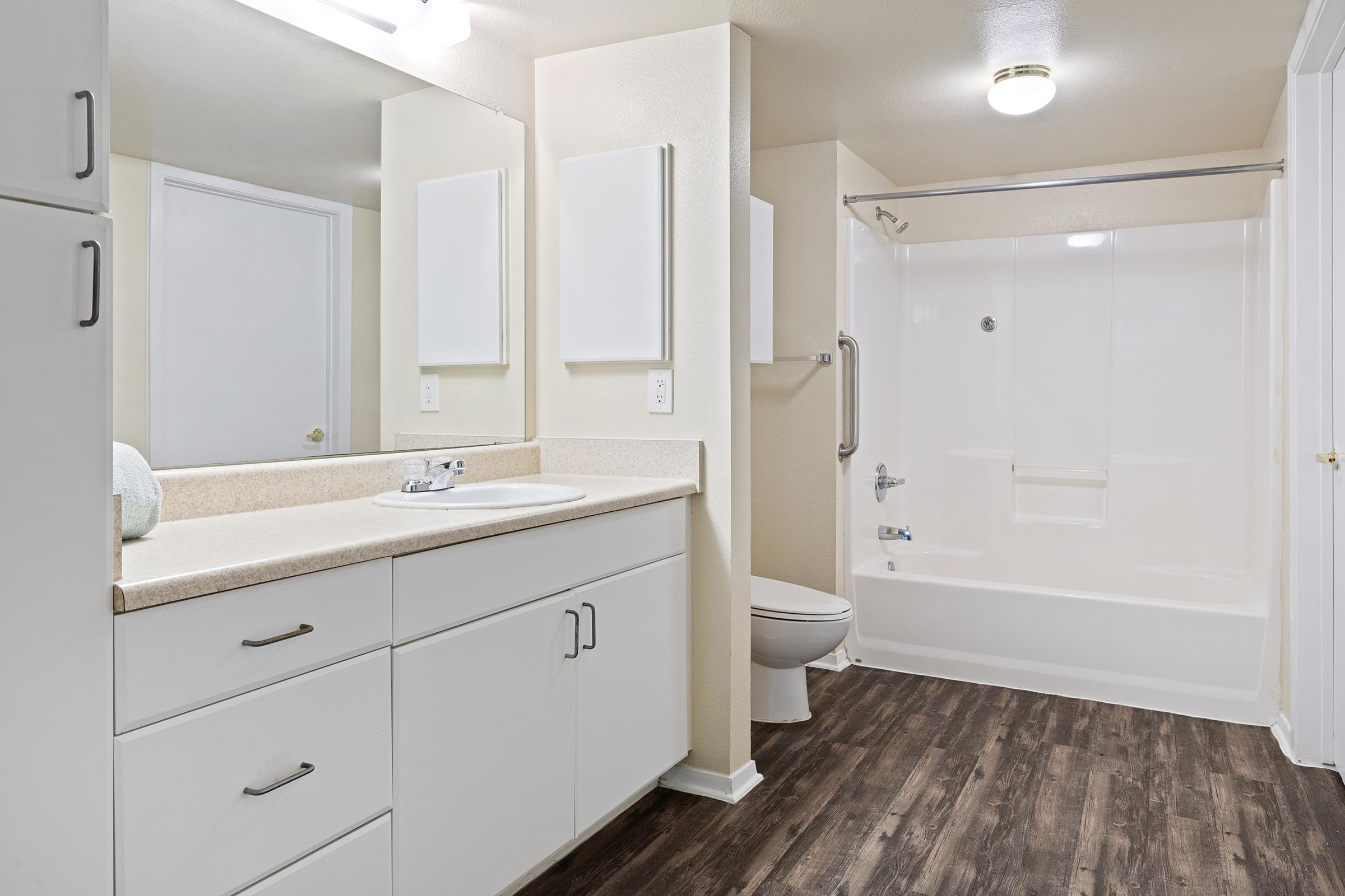 A modern bathroom featuring white cabinets and a countertop, a large mirror above the sink, a toilet, and a bathtub with an overhead shower. The walls are light-colored, and the flooring is dark wood laminate. Soft lighting enhances the clean, contemporary look of the space.