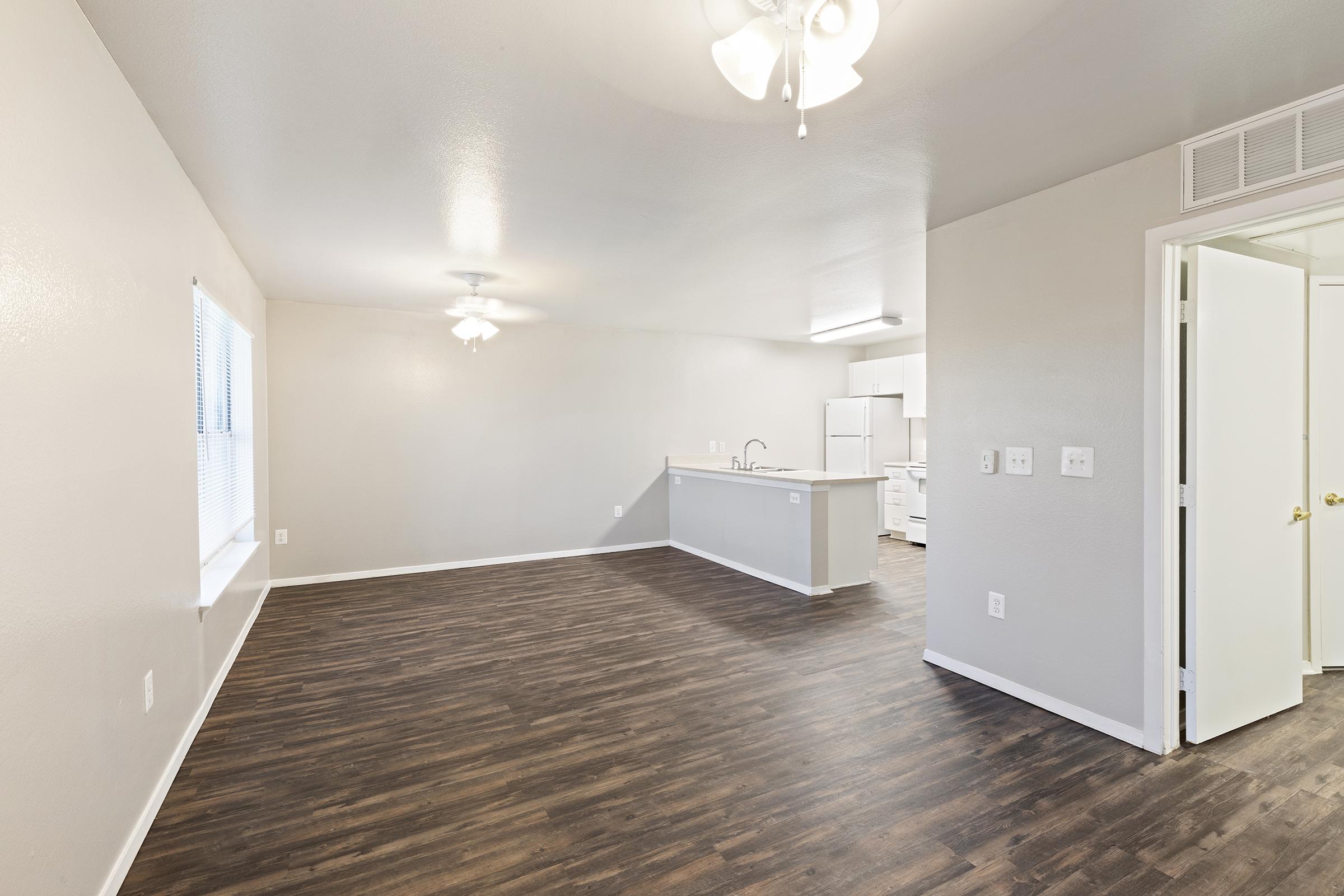 A spacious, well-lit interior of an empty living room with hardwood flooring. It features a ceiling fan, white walls, and an open layout leading to a kitchen area with modern appliances. A door leads to another room, and there is a large window allowing natural light.