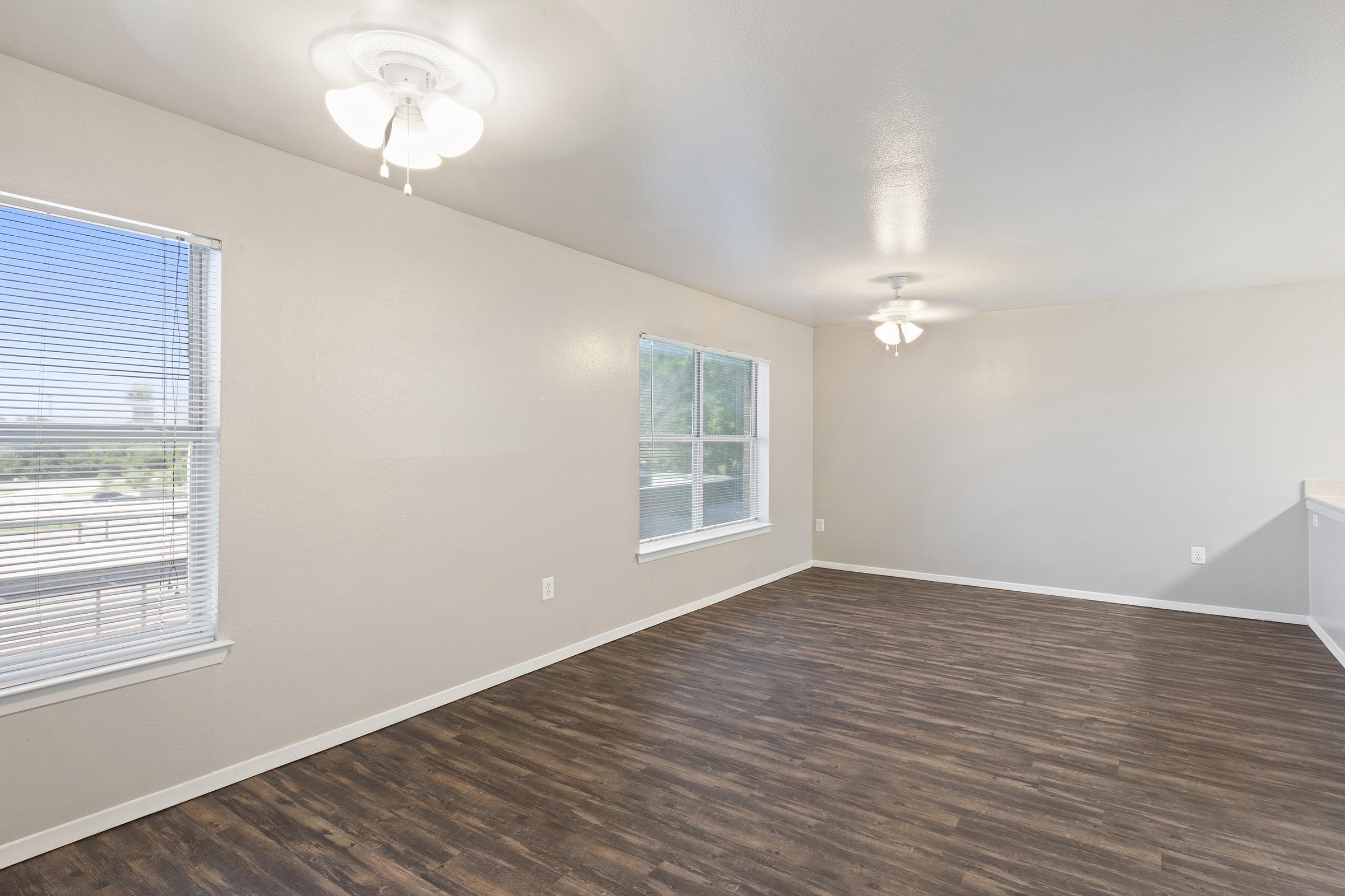 Empty room with light-colored walls and wood laminate flooring. Two ceiling fans are visible, and there are two windows with horizontal blinds allowing natural light to enter. The room appears well-lit and spacious, ideal for various uses.