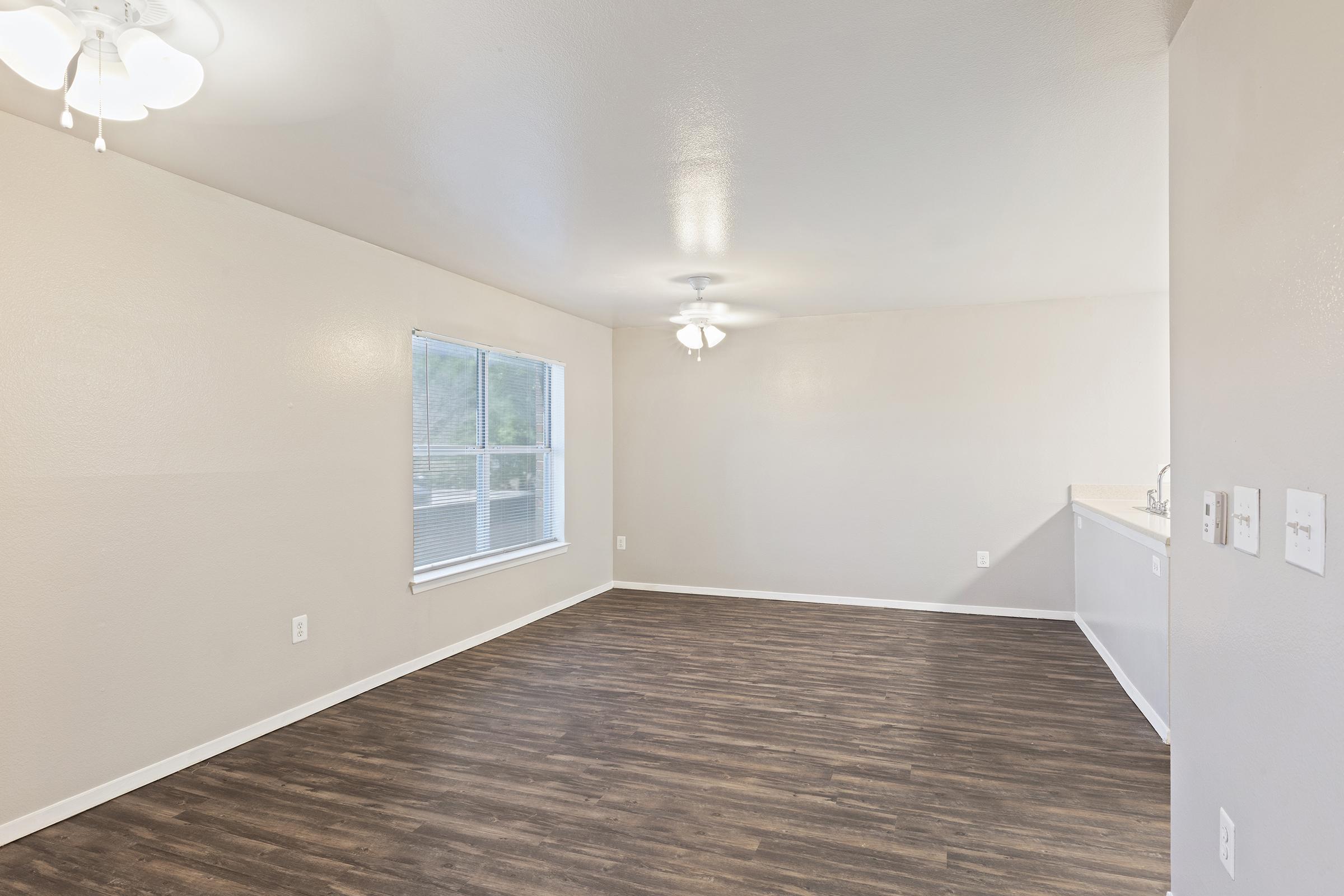 Spacious, empty room with light-colored walls and a large window. The floor is made of dark, wood-like material. A ceiling fan hangs in the center, and there's a glimpse of a kitchen area in the background. Natural light brightens the space, creating a welcoming atmosphere.