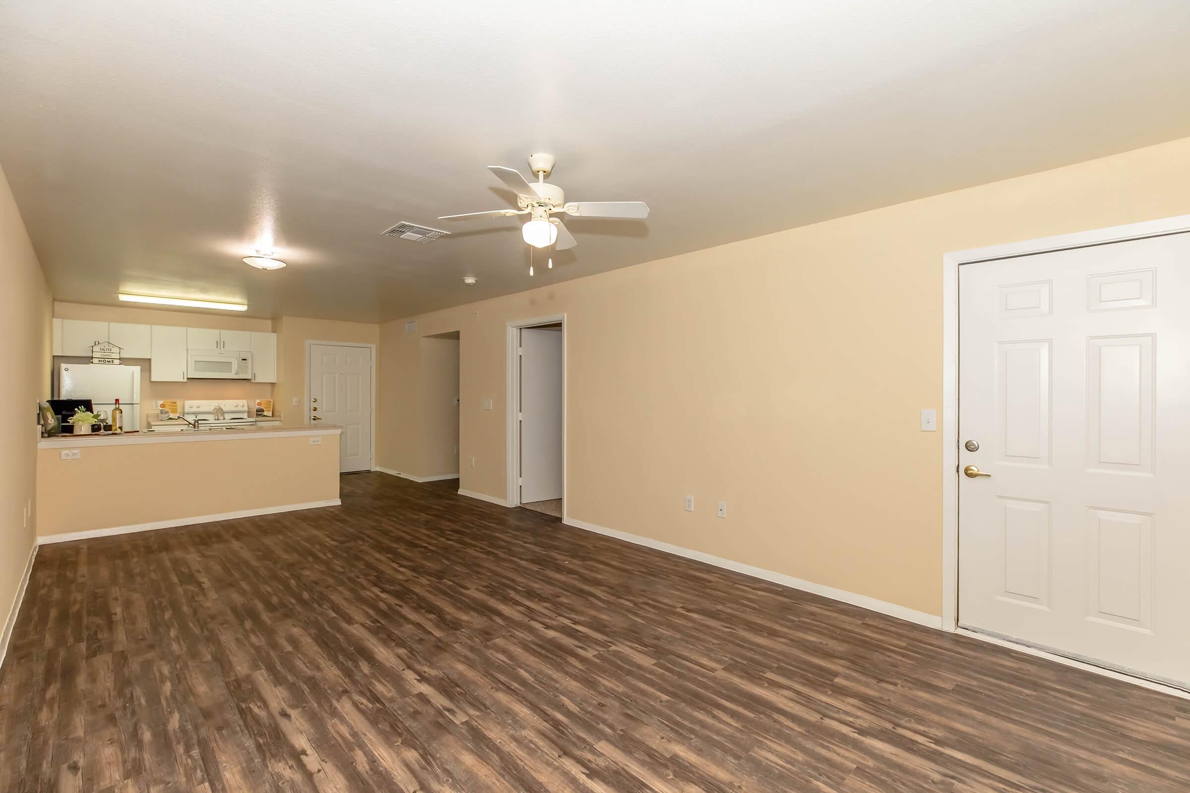 Spacious interior of an apartment featuring light-colored walls, a ceiling fan, and a door leading outside. The living area has laminate flooring, with an open kitchen view in the background. The kitchen is partially visible, showcasing white appliances and cabinets.