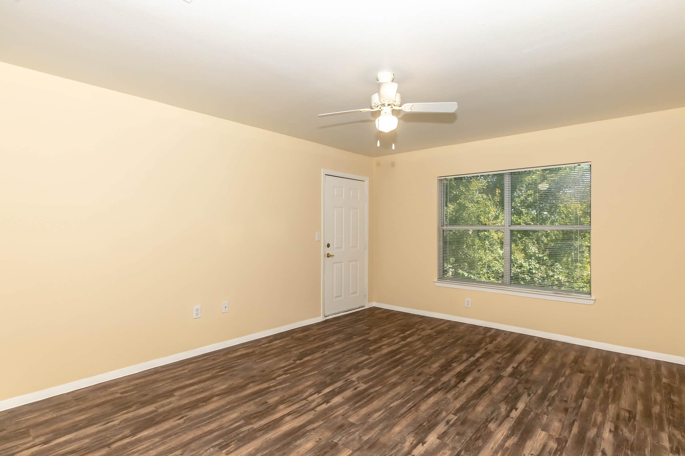 A spacious, empty room with light beige walls and a ceiling fan. It features a white door and a large window providing natural light, revealing greenery outside. The floor is covered with wooden laminate, enhancing the room's warmth and openness.
