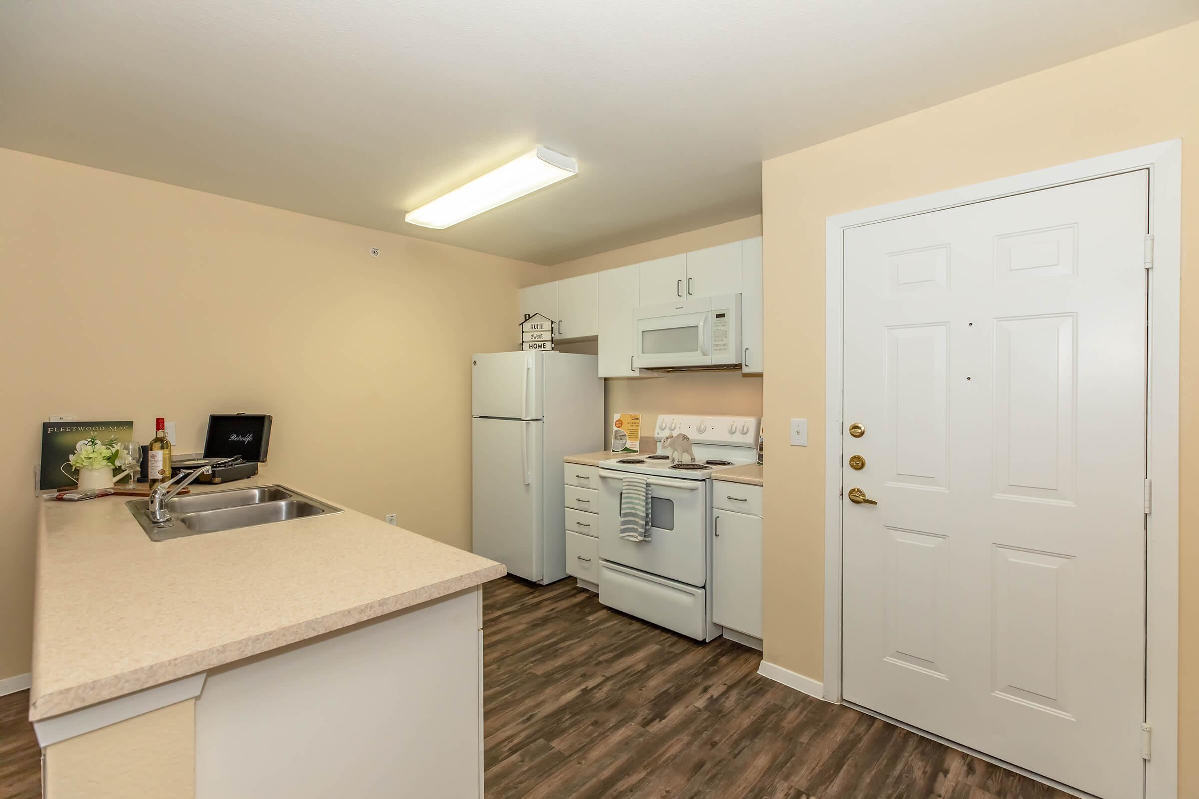 A modern kitchen featuring white cabinets, a refrigerator, a microwave, and an oven. The countertop is light-colored with a small sink. The walls are painted a soft beige, and there's a front door visible with a doorknob. Natural light from an overhead fixture brightens the space.