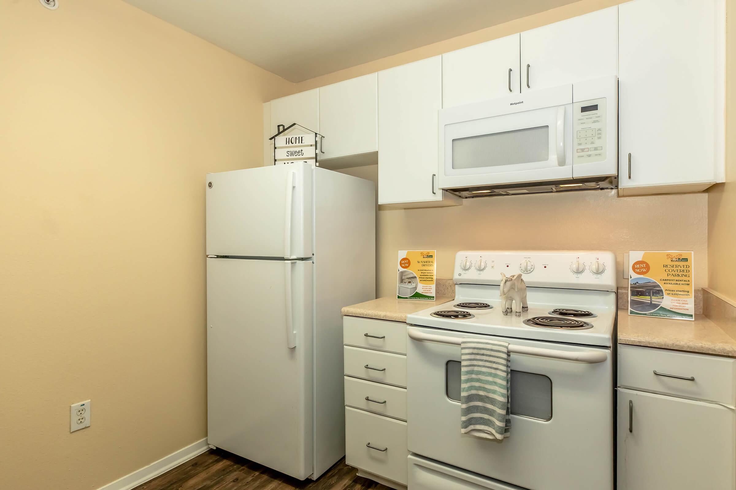 A modern kitchen featuring a white refrigerator, microwave, and stove with an oven. The countertop is light-colored, and a striped dish towel hangs from the stove. Two informational signs are placed on the counter, and the walls are painted a warm beige. The flooring is a wooden design.