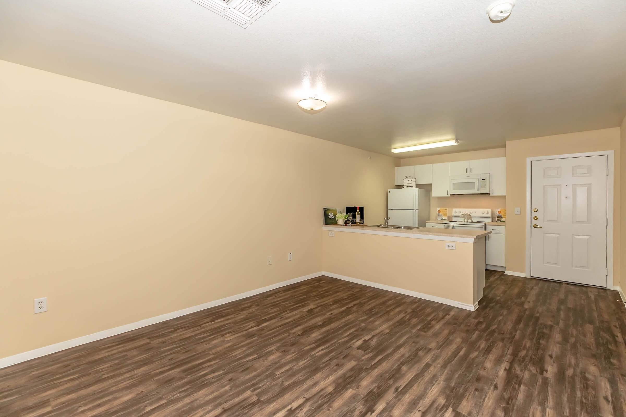A spacious, empty apartment interior featuring a light beige wall color and large windows. The kitchen area is visible with white cabinetry and appliances, while the living area has hardwood-style flooring. The front door is on the right, and there's a ceiling light fixture providing illumination.