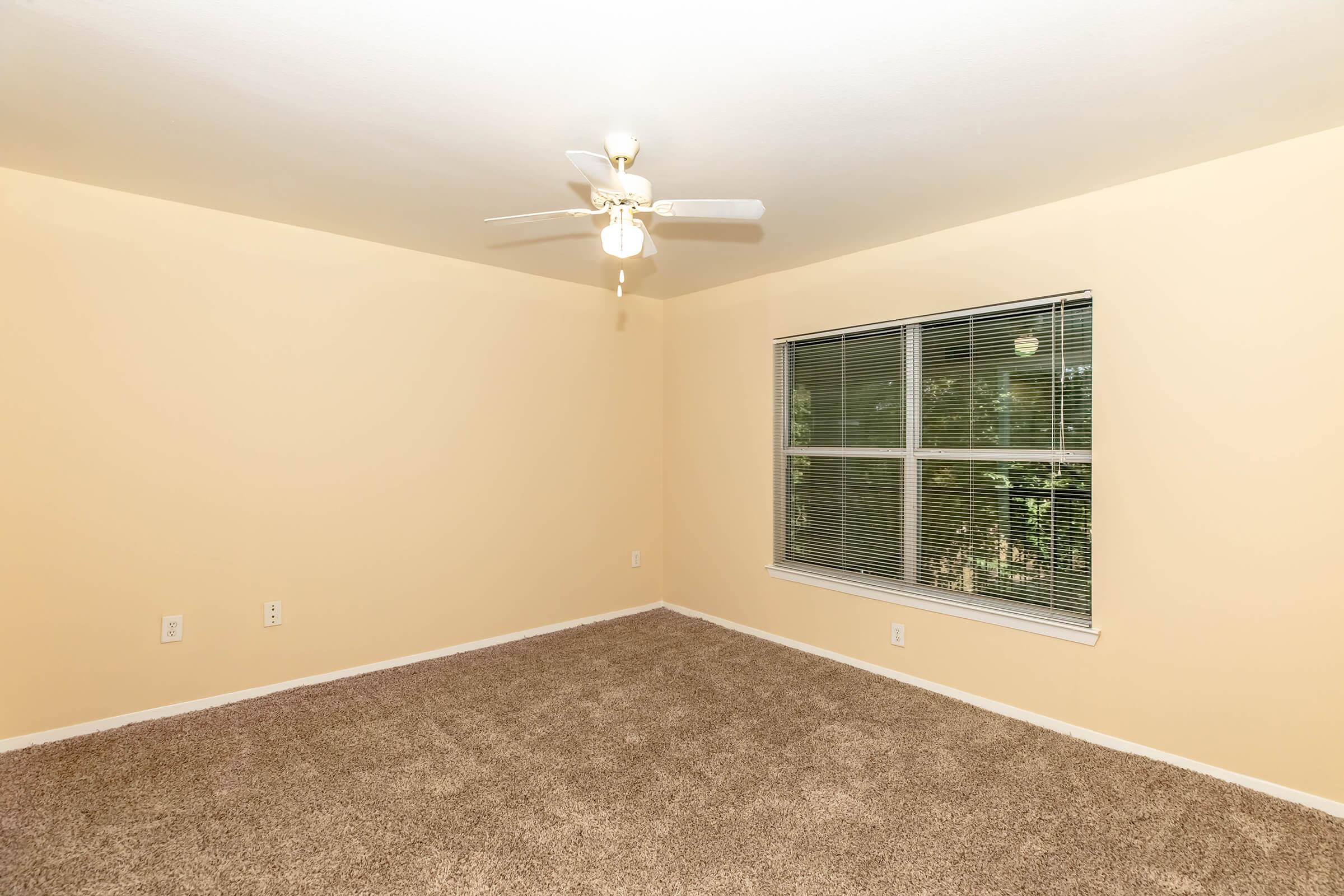 A spacious, empty room with light beige walls and plush carpet flooring. A ceiling fan hangs from the ceiling, providing a modern touch. A large window with multiple panes allows natural light to enter, showcasing an outdoor view with greenery. The room appears clean and ready for furnishing.