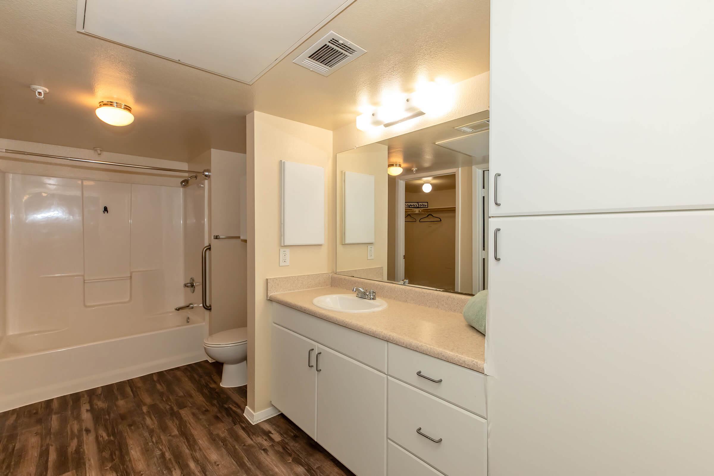 A clean and modern bathroom featuring a white bathtub and shower, a toilet, and a double vanity with a mirror. The cabinetry is light-colored, and the flooring is a dark wood style. There are two wall-mounted mirrors and a soft green towel on the counter, creating a welcoming atmosphere.