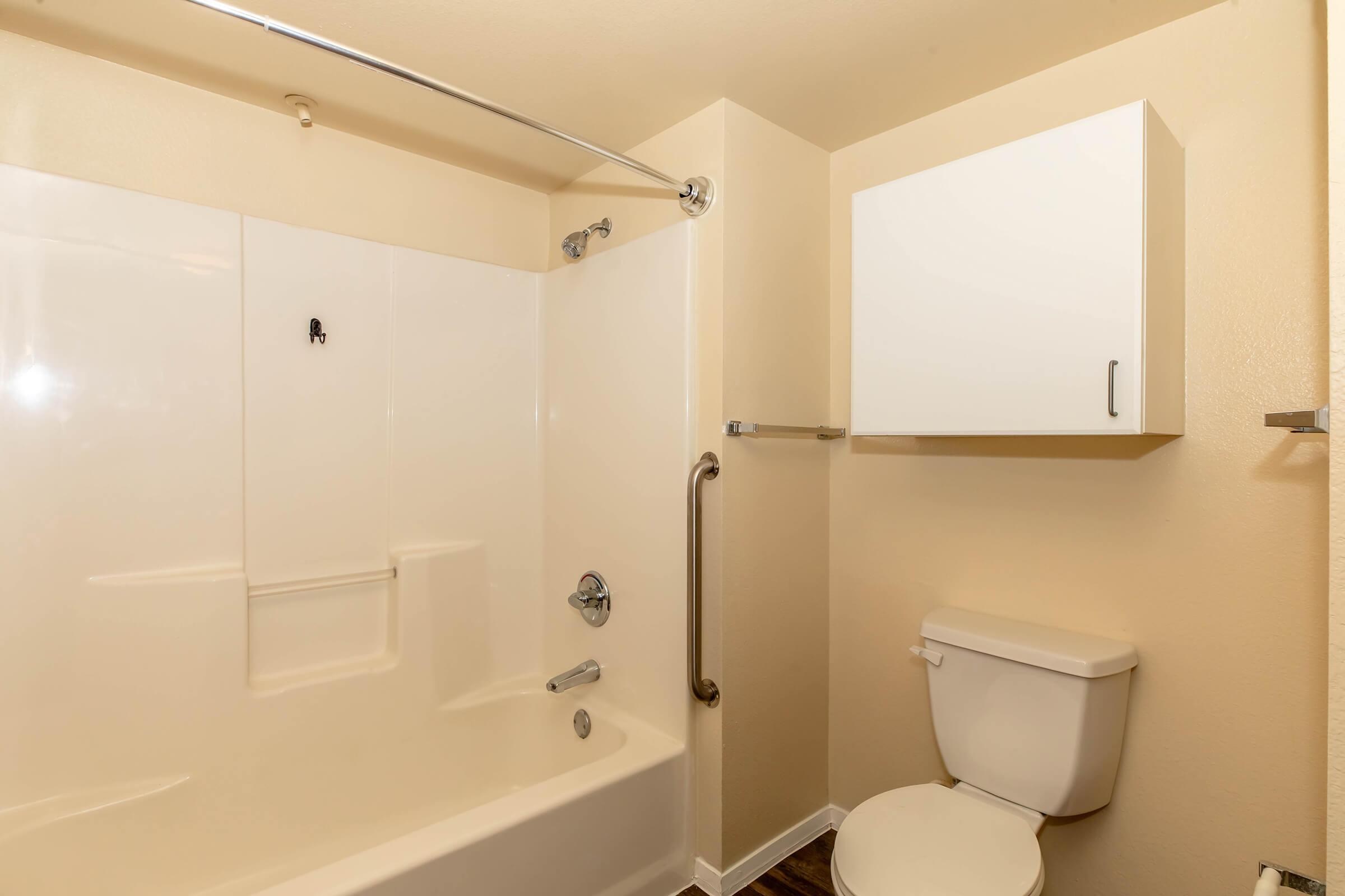 A simple and clean bathroom featuring a white bathtub with a shower, a toilet, and a mounted cabinet. The walls are painted light beige, and there's a grab bar for safety. The flooring is wood-like. The overall aesthetic is modern and functional.