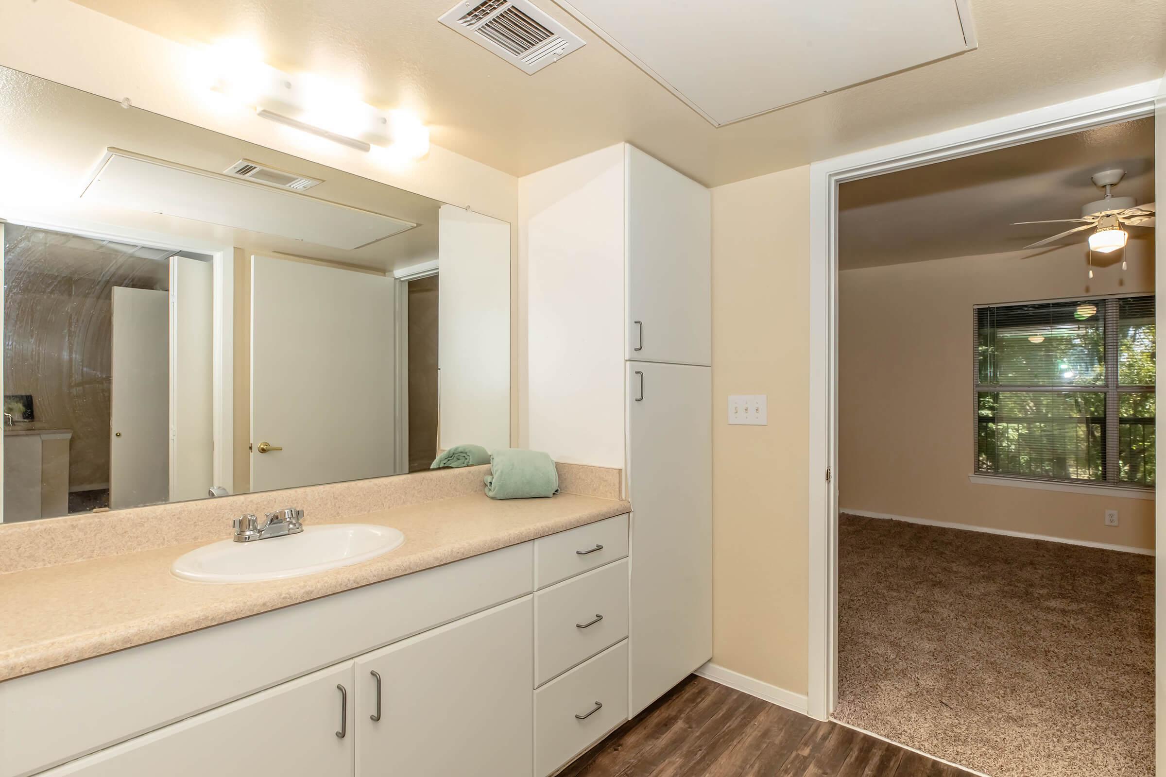 A well-lit bathroom featuring a long counter with a sink and mirror, white cabinetry, and a green towel. The doorway leads to a carpeted room with a window, allowing natural light to enter. The overall color scheme is neutral, with light-colored walls and flooring.