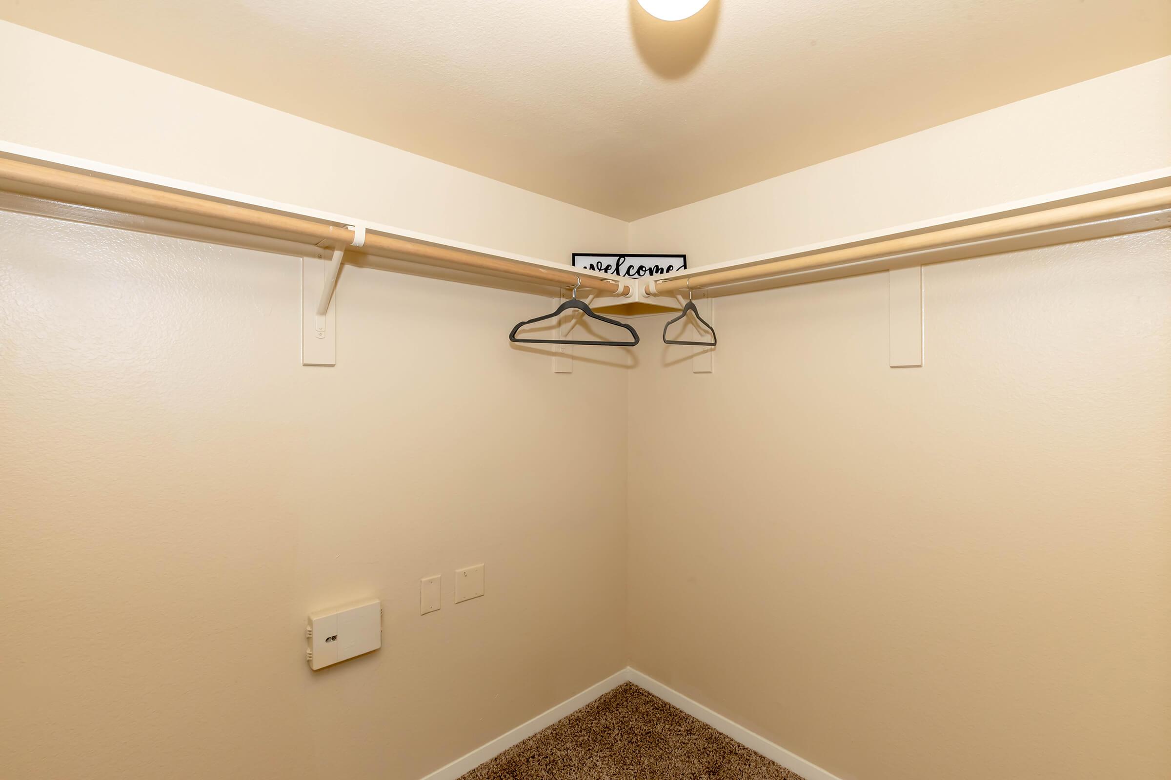 Empty closet with beige walls and carpeted floor. Two hanging rods with a few empty hangers and a small sign that says "welcome" on a shelf above. There are electrical outlets on the wall, but no additional items or furnishings are present. Soft lighting from a ceiling fixture.