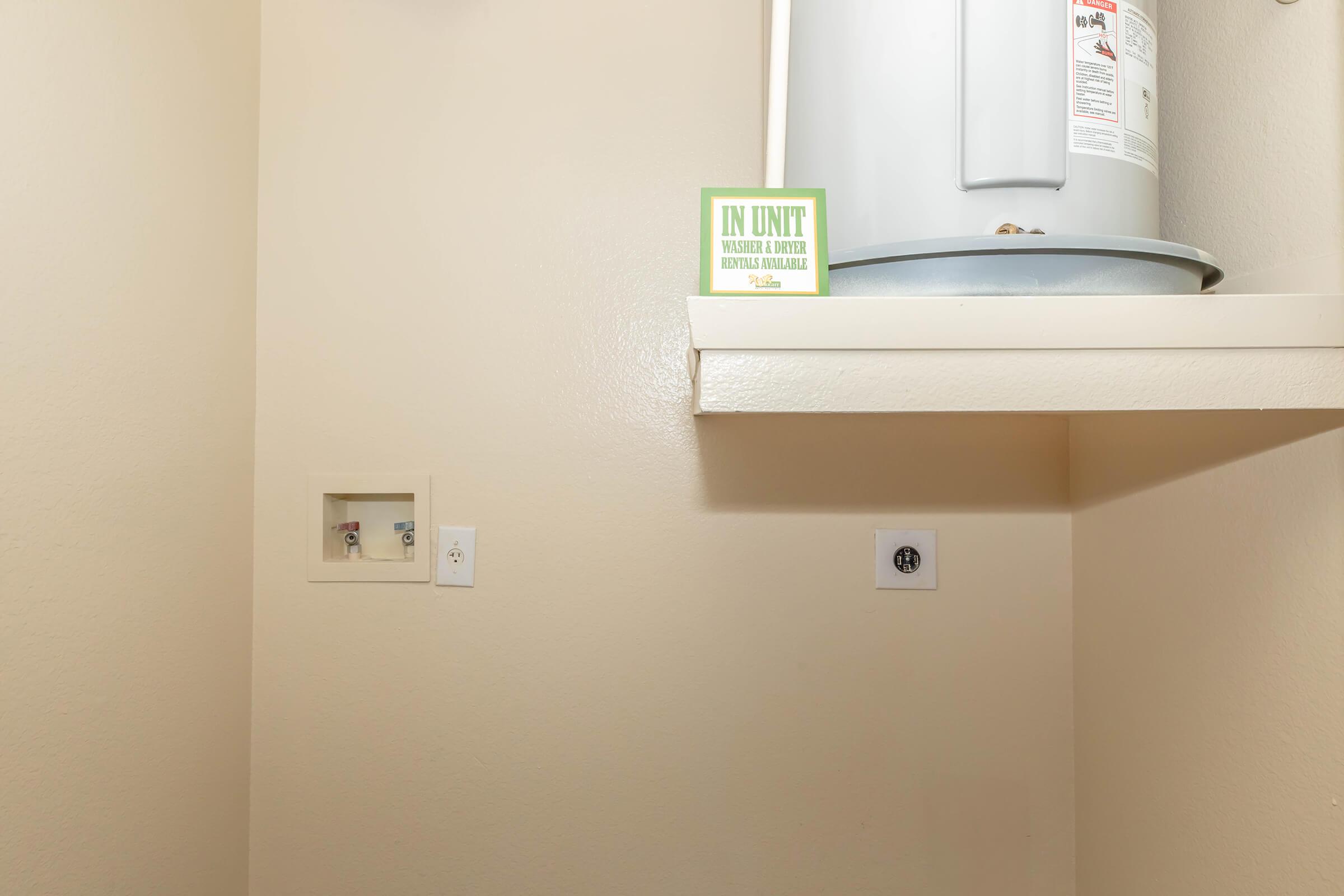 A small, light-colored utility room with a water heater on a shelf. There are two electrical outlets on the wall and a notice sign that reads "IN UNIT" indicating that water and dryer hookups are available. The overall space appears clean and minimalistic.