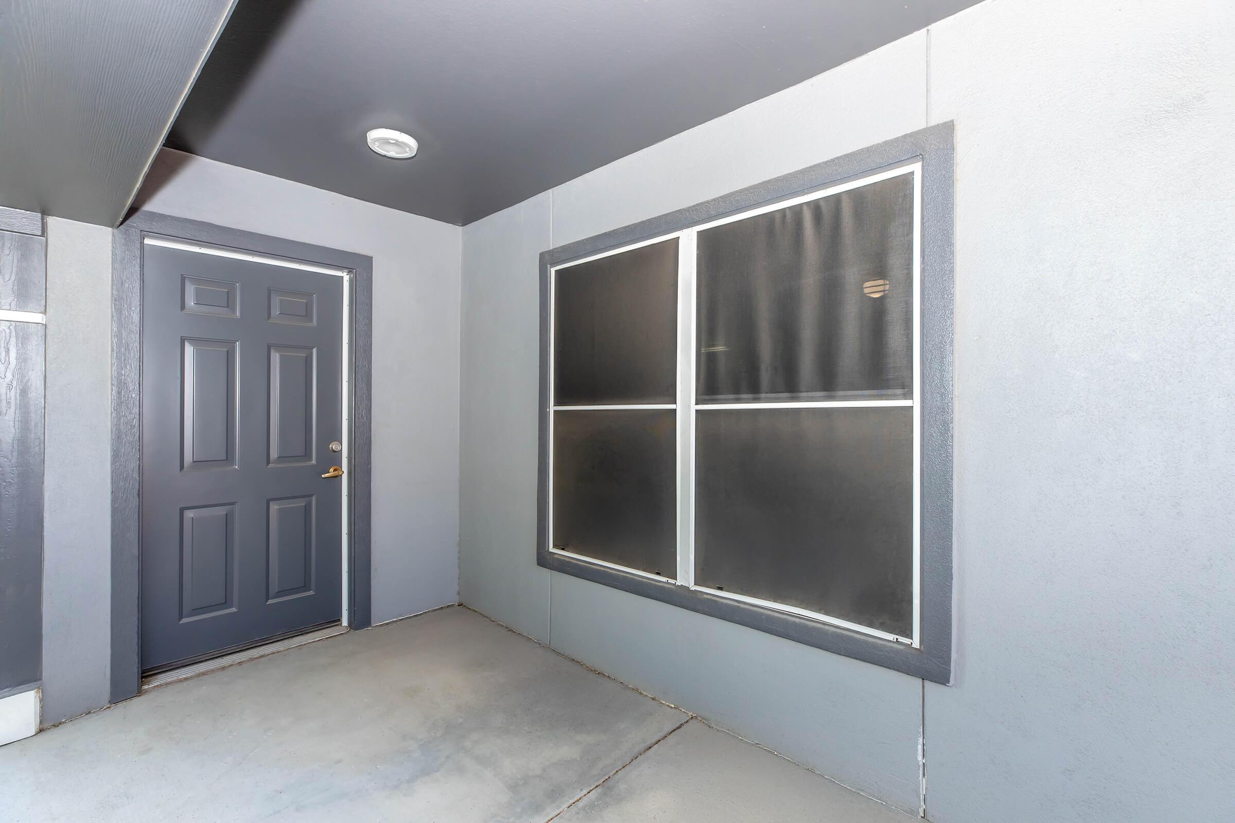 A gray exterior entrance with a dark front door and a large window covered by a black screen. The walls of the entrance are also gray, and the ground is concrete. The setting is simple and modern, creating a clean and minimalistic atmosphere.
