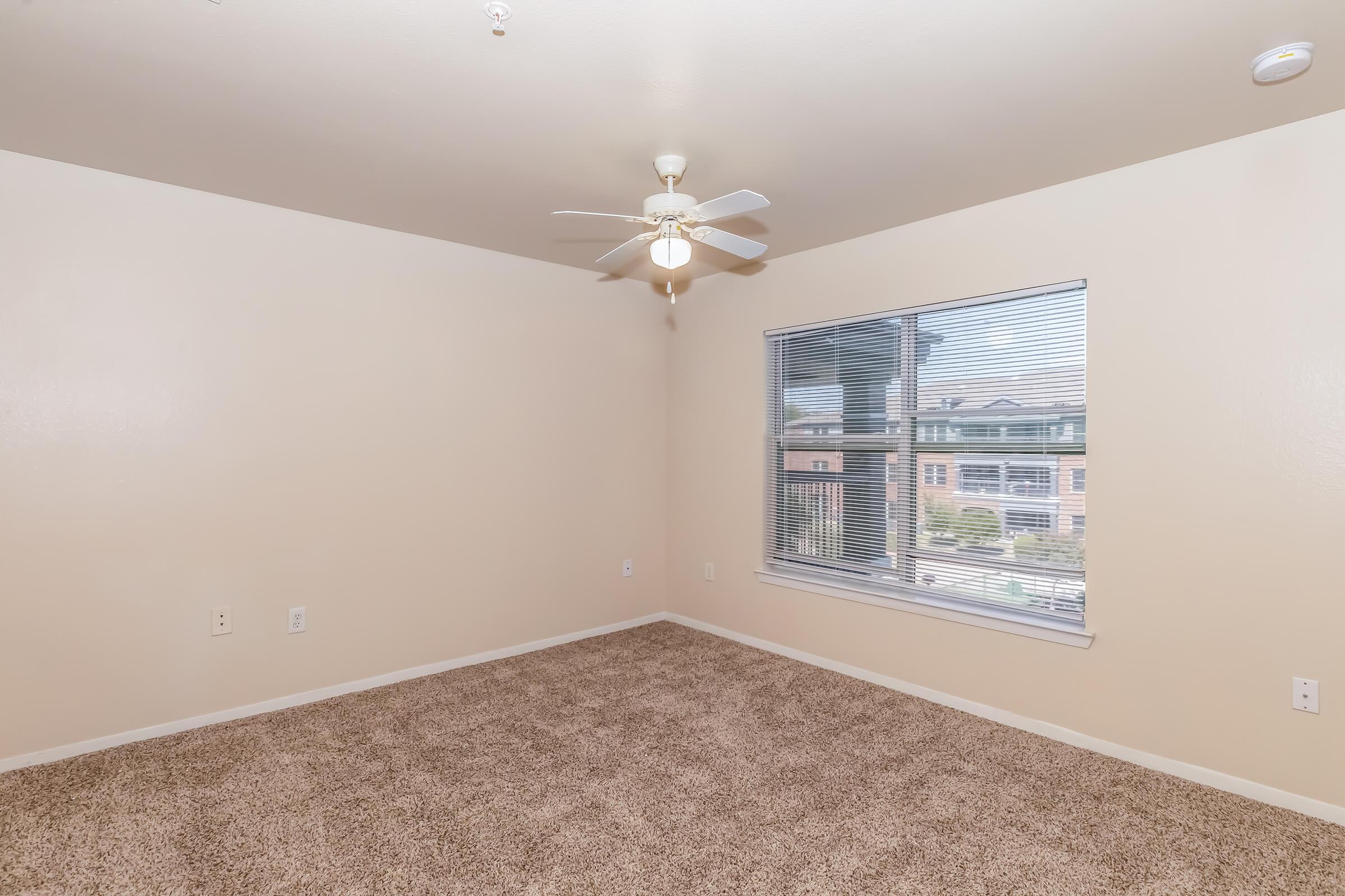 Empty room with light beige walls and carpeted floor. A ceiling fan is mounted overhead. A large window with blinds allows natural light to enter, revealing a view of an outdoor area. The space is unfurnished, creating a blank canvas for interior design.