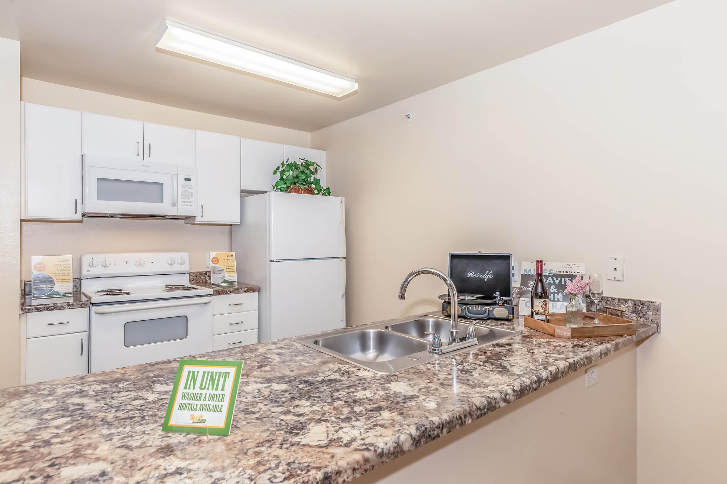 A modern kitchen featuring white appliances, including a microwave and refrigerator, on light-colored cabinets. The countertop is made of speckled granite. There are decorative items and a small potted plant. A sign reading "IN UNIT" is placed on the counter.