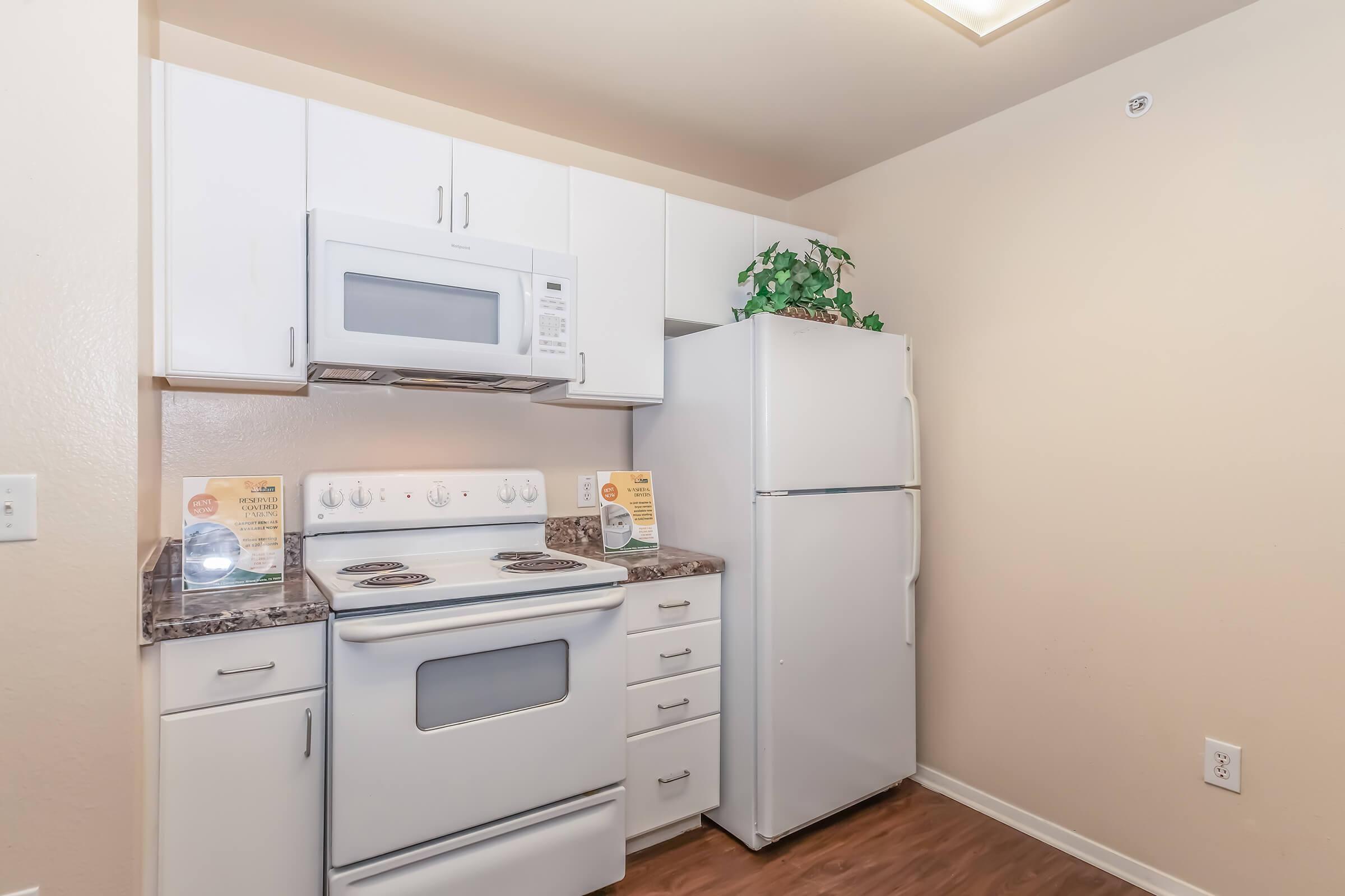 A small kitchen featuring a white stove and microwave, a white refrigerator, and light-colored cabinets. The countertop has a dark finish and is adorned with potted greenery. Informational signs are placed on the counter, providing details about the kitchen appliances. The walls are painted in a neutral color.