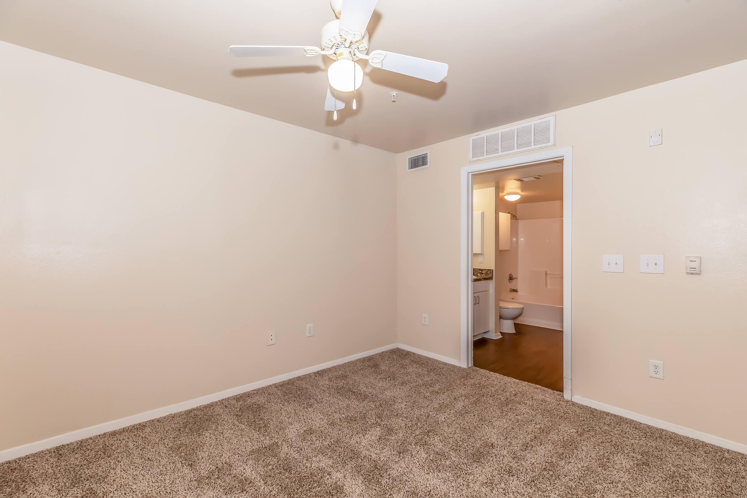 A spacious room with light beige walls and a ceiling fan. The floor is covered in plush beige carpet. In the background, a doorway leads to a bathroom area with light fixtures visible. The overall ambiance is warm and inviting, suitable for various interior designs.
