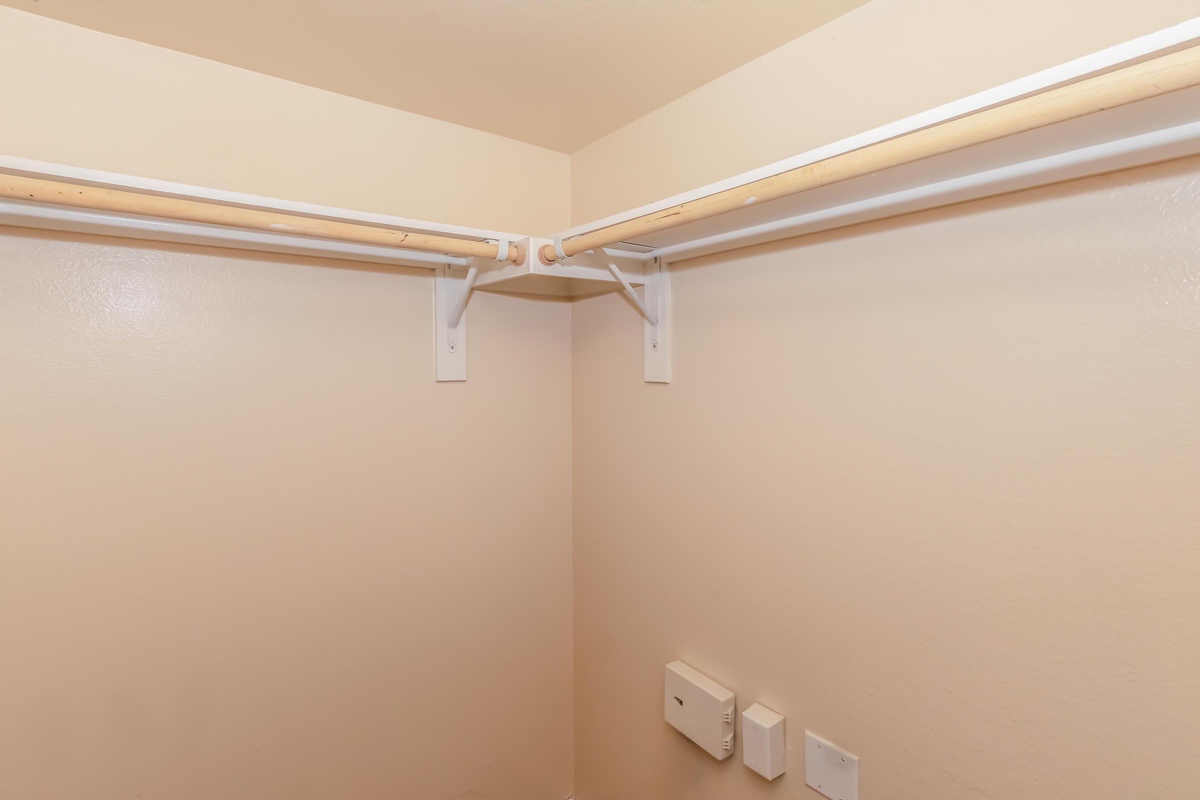 Empty corner of a closet with two hanging rods mounted on white walls. There are two electrical panels or switches on the wall, and the floor is bare. The overall color scheme is neutral with light beige tones.