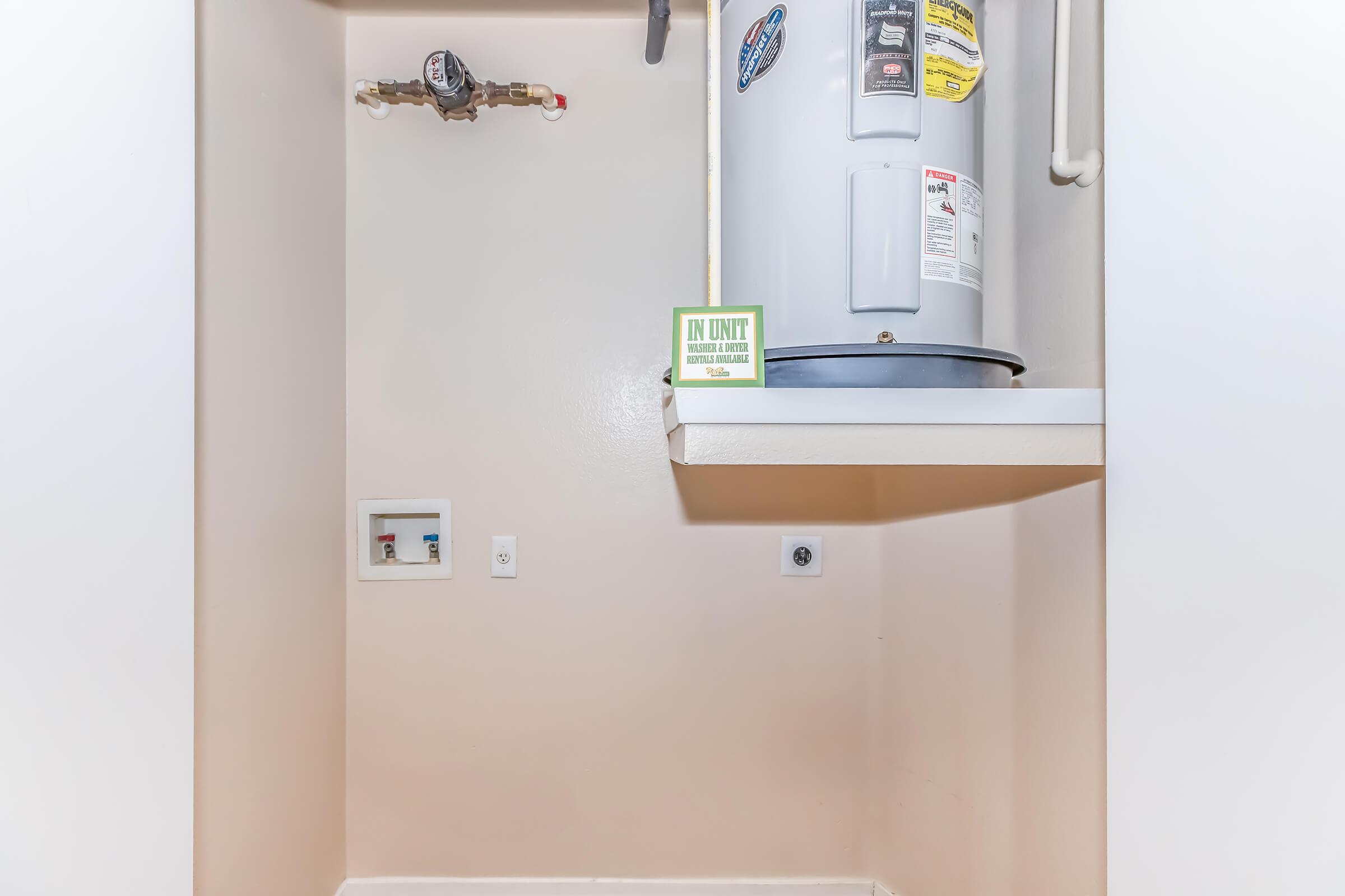 A small utility closet featuring a water heater mounted on the wall, with a green sign labeled "IN UNIT" displayed on a shelf. There are water supply pipes visible, along with electrical outlets and controls on the wall. The environment is clean and well-lit, with light-colored walls.