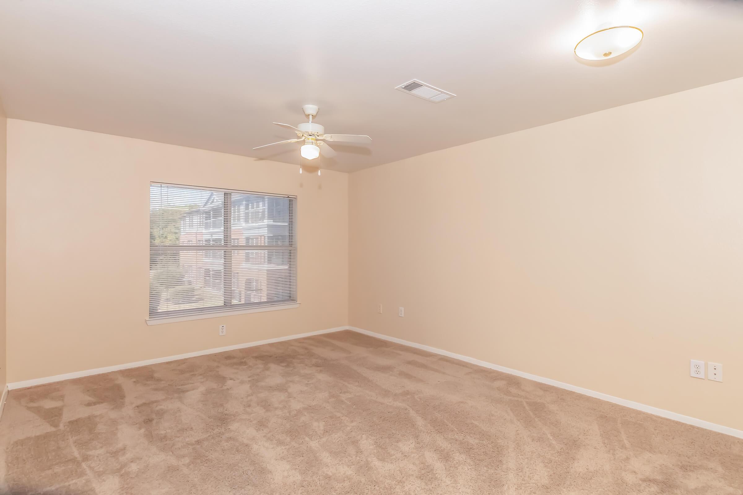 Empty room featuring light beige walls and carpeted flooring. A ceiling fan is installed, and there's a window allowing natural light, revealing an exterior view. The space is well-lit with an overhead light fixture, creating a neutral and inviting atmosphere.