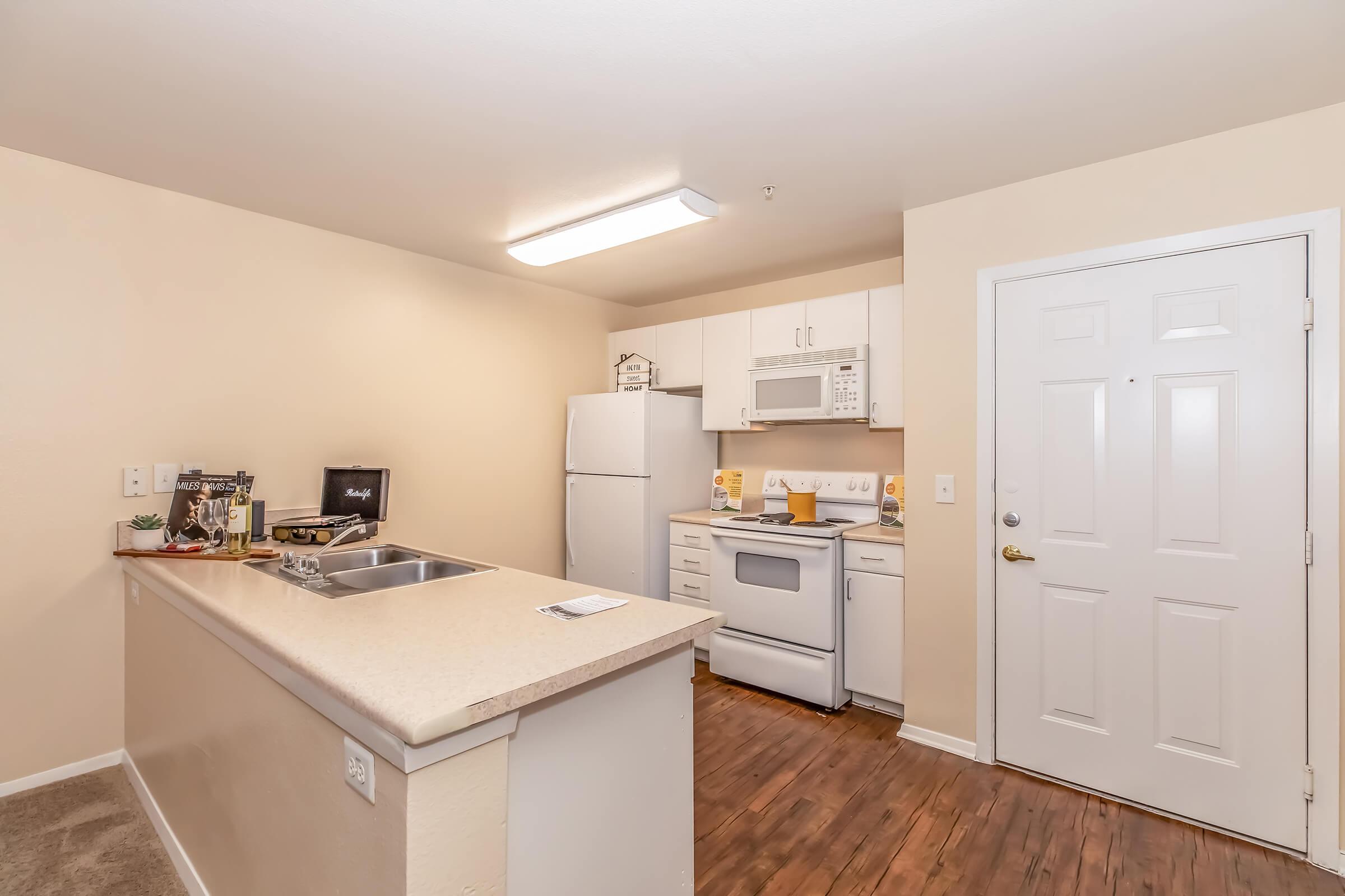 A modern kitchen featuring white cabinets, a refrigerator, microwave, and stove. The countertop offers a sink and some decorative items. The walls are painted a light color, and there's a door leading outside. The flooring is wood-like, adding warmth to the space.
