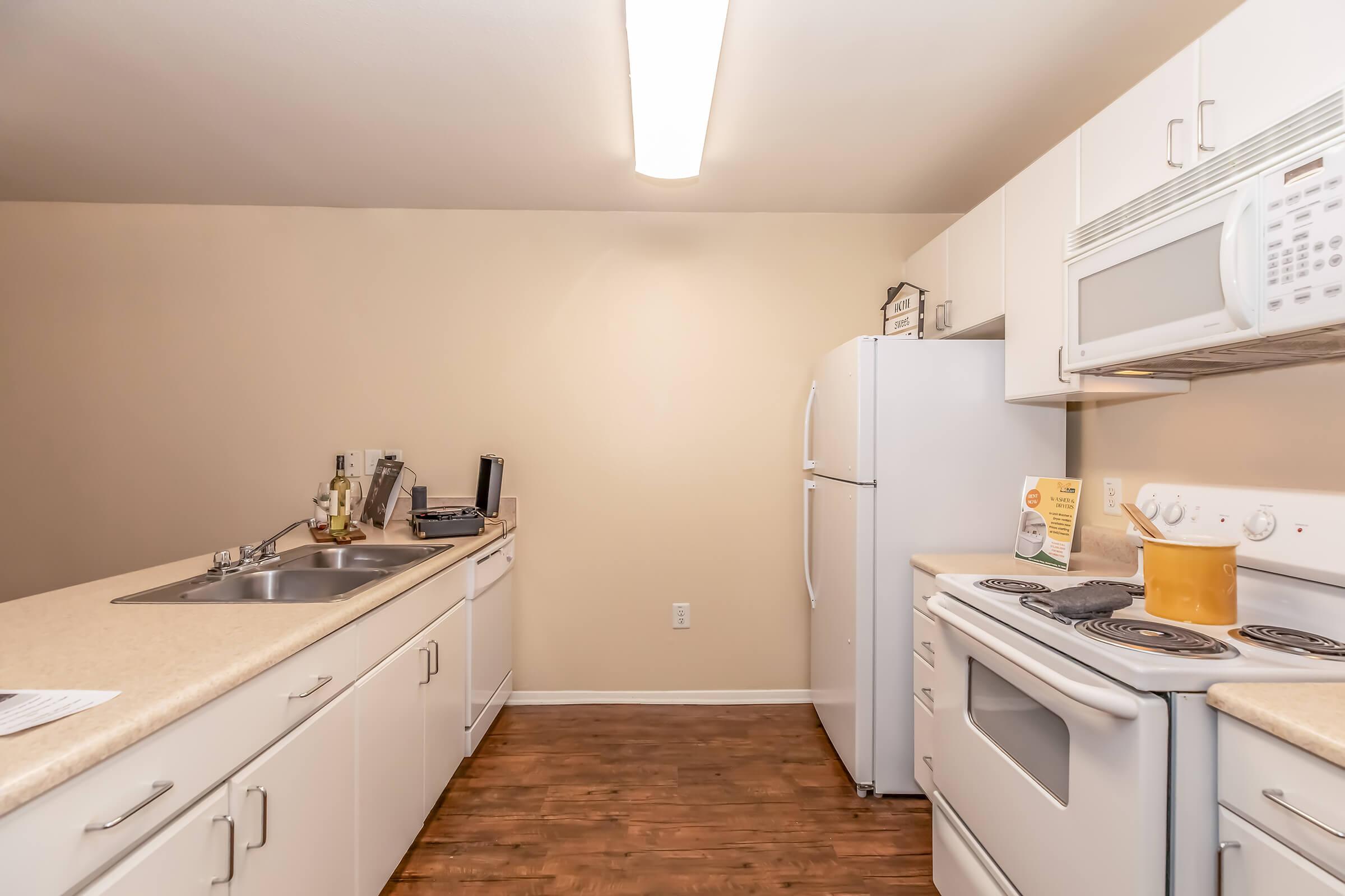 A modern kitchen featuring white cabinetry, a double sink, a refrigerator, a microwave, and a stove. The countertops are light-colored, and the floor is made of dark wood. The walls are painted a soft beige, creating a bright and inviting atmosphere. A small window may provide natural light.
