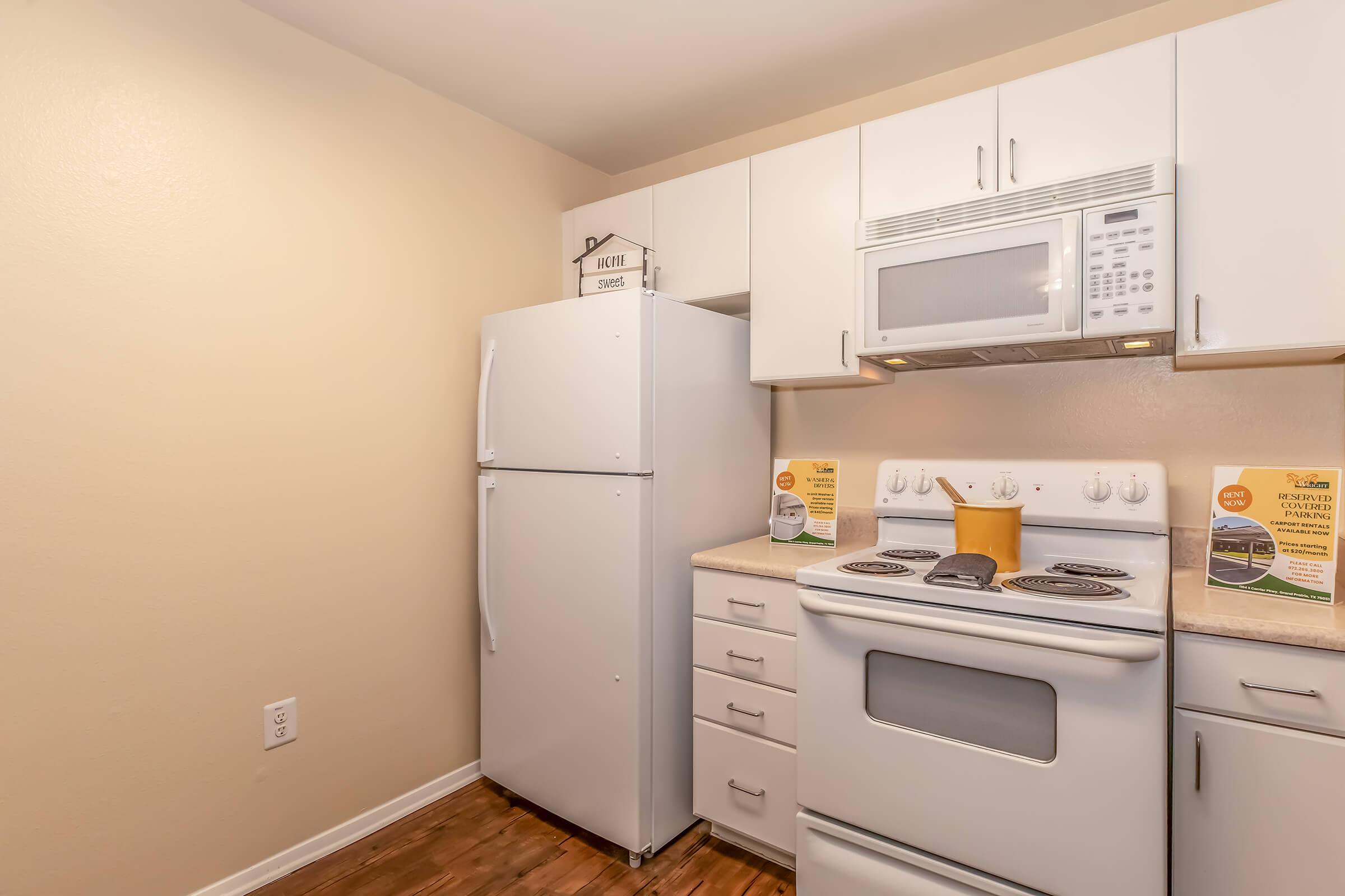 A modern kitchen featuring a white refrigerator, a built-in microwave above an electric stove, and white cabinets. The countertop has a yellow cooking pot, and there are informational flyers displayed on the counter. The walls are painted a light beige, and the flooring is a warm wood finish.