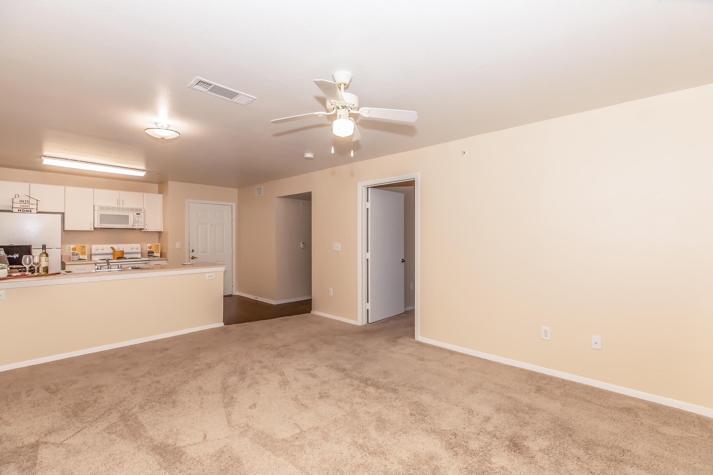 Spacious living area in a light-colored apartment with beige carpet. The kitchen is visible in the background, featuring white cabinets and appliances. A ceiling fan hangs above, and there are two doorways leading to other rooms. Walls are painted in a neutral tone, creating a bright and inviting atmosphere.